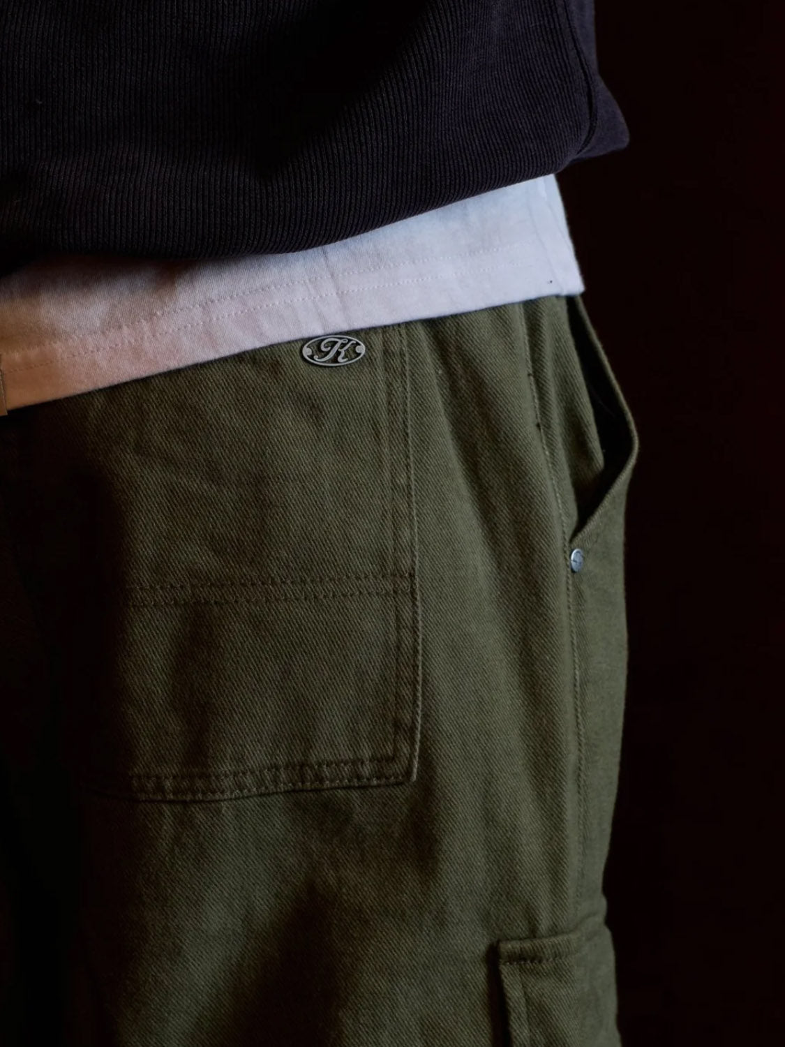 A close-up of a person wearing Kuwalla’s Cargo Trouser in relaxed fit olive green cotton, featuring a patch pocket and small metal logo, styled with a white t-shirt and dark jacket. The focus is on the waist and hip area.