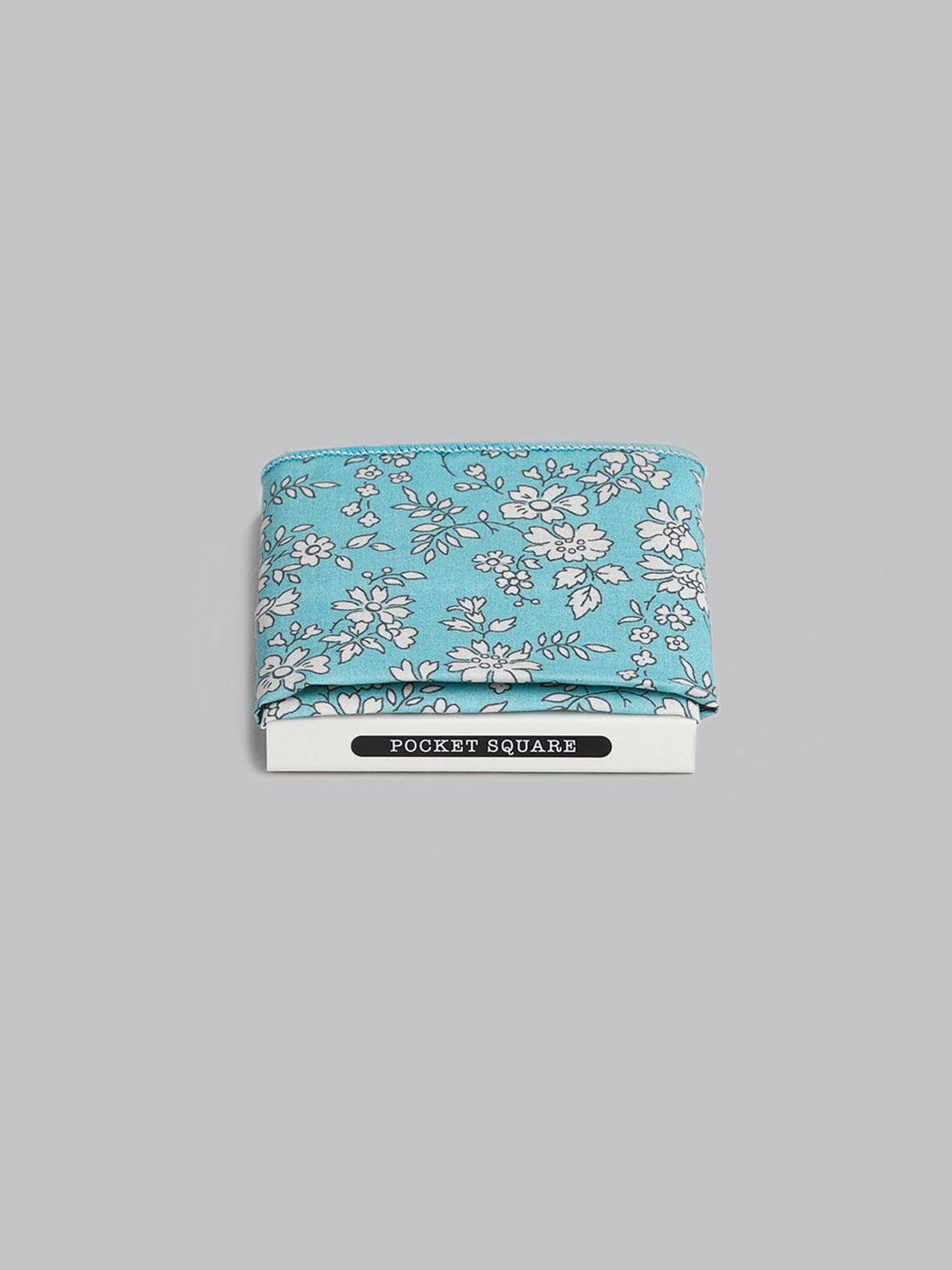 The Liberty™ Carpel Pocket Square by ID Menswear, handmade in Brooklyn from blue floral Tana Lawn cotton, is displayed atop packaging marked POCKET SQUARE against a plain gray background.
