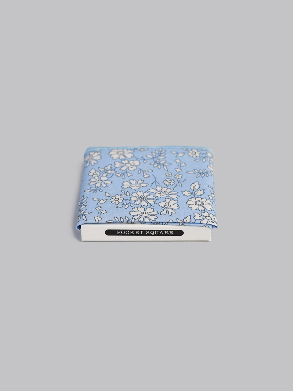 The Liberty™ Carpel Pocket Square by ID Menswear, handmade in Brooklyn from light blue Tana Lawn cotton with a white floral pattern, is shown partially tucked into a card labeled POCKET SQUARE on a plain gray background.
