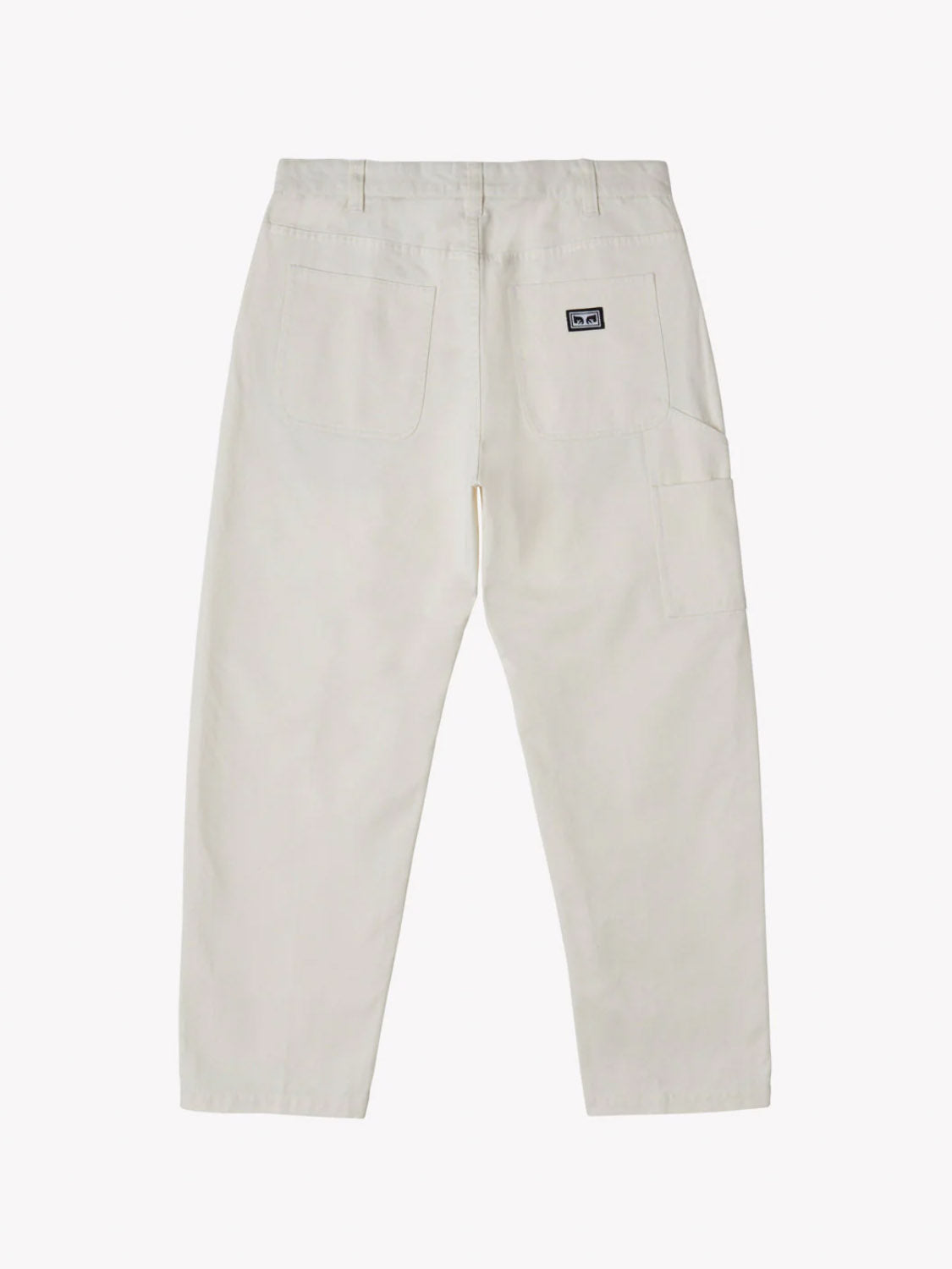 Back view of Obey Hardwork Carpenter Pant in light beige twill, showcasing two large carpenter pockets, a small logo patch on the right pocket, and a lower side pocket on the left leg. These Obey pants have a regular fit and straight cut, set against a plain white background.