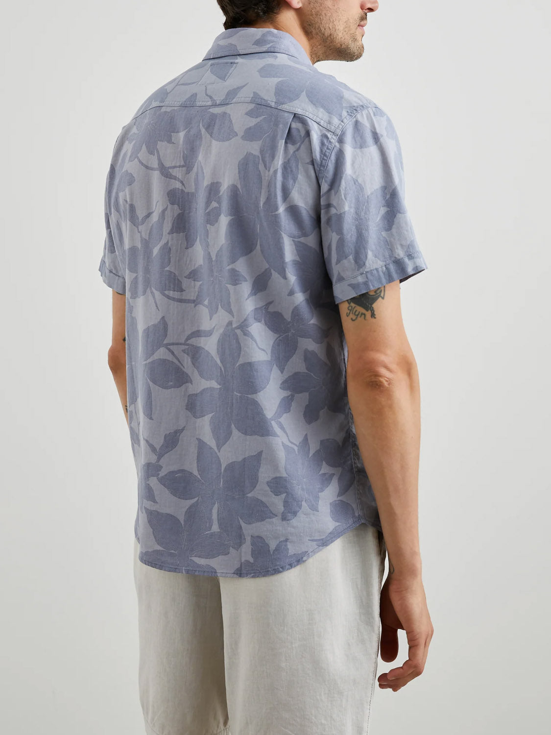A person wears the Rails Carson Garden Sands Orchid, a lightweight linen-blend short-sleeve button-down in light blue with a subtle leaf pattern. Facing away, they pair it with light-colored shorts and display an arm tattoo against a minimalistic backdrop.