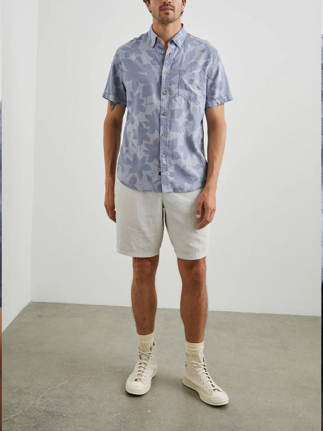 A person wearing the Rails Carson Garden Sands Orchid shirt and lightweight linen blend shorts stands in a minimalist room, sporting cream-colored high-top sneakers and light socks against a plain light gray wall and floor backdrop.