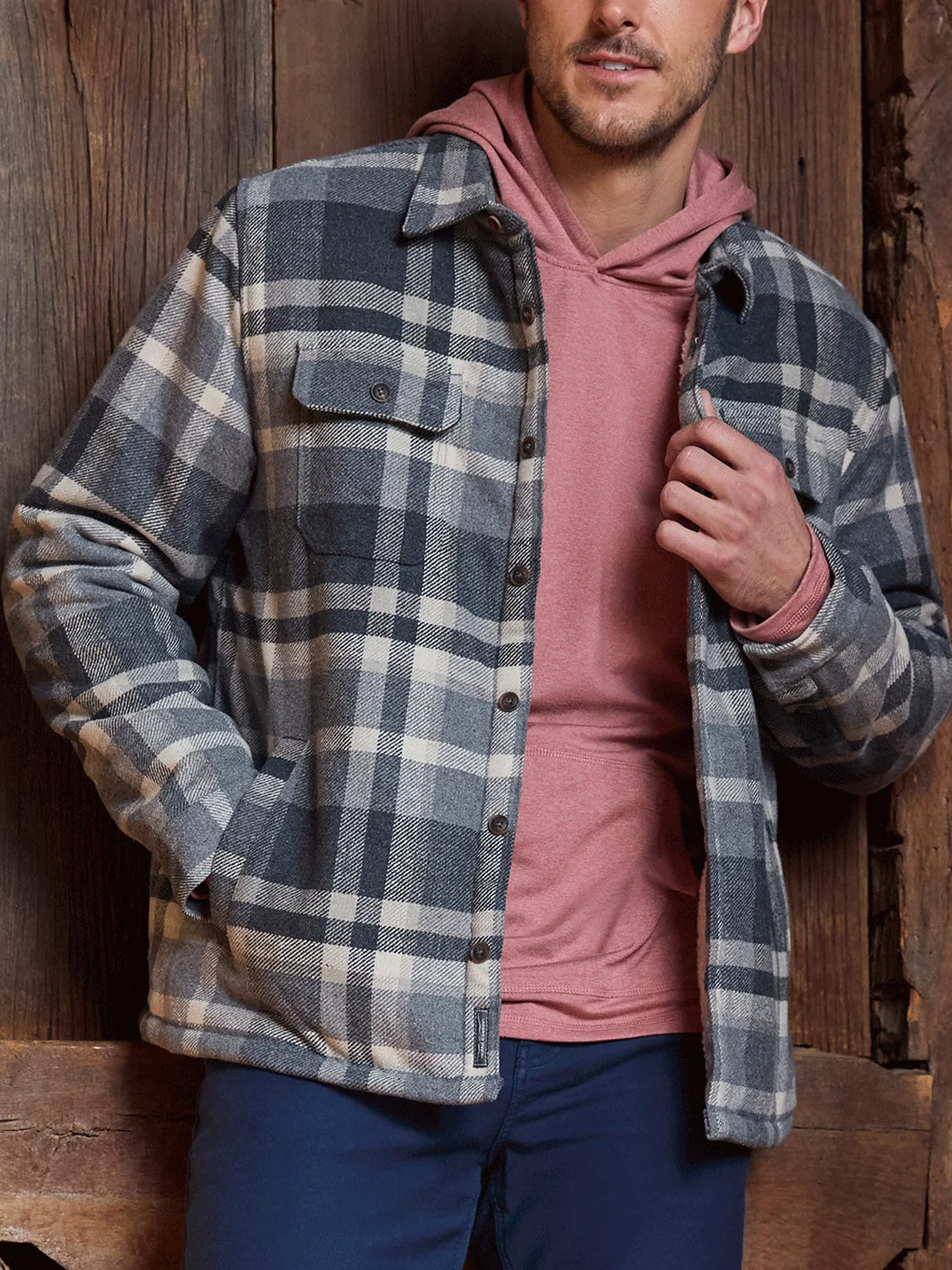 A man stands against a wooden wall, wearing the Flag and Anthem Cassidy Sherpa Lined Shirt Jacket in gray and white plaid over a pink hoodie and dark pants, one hand in his pocket, the other adjusting his jacket.