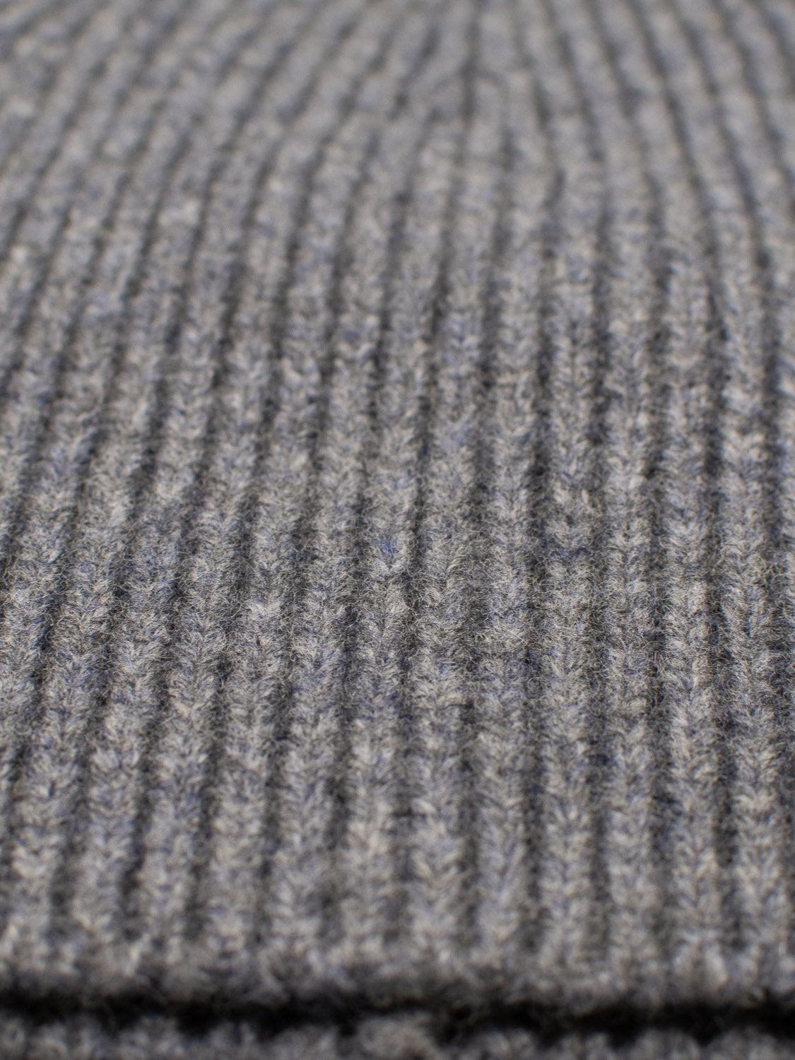 Close-up of a knitted gray fabric with ribbed texture, featuring parallel lines creating a pattern across the 100% wool material. It highlights the soft and warm appearance typical of winter wear, like ID Menswears Chamonix Wool Beanie.