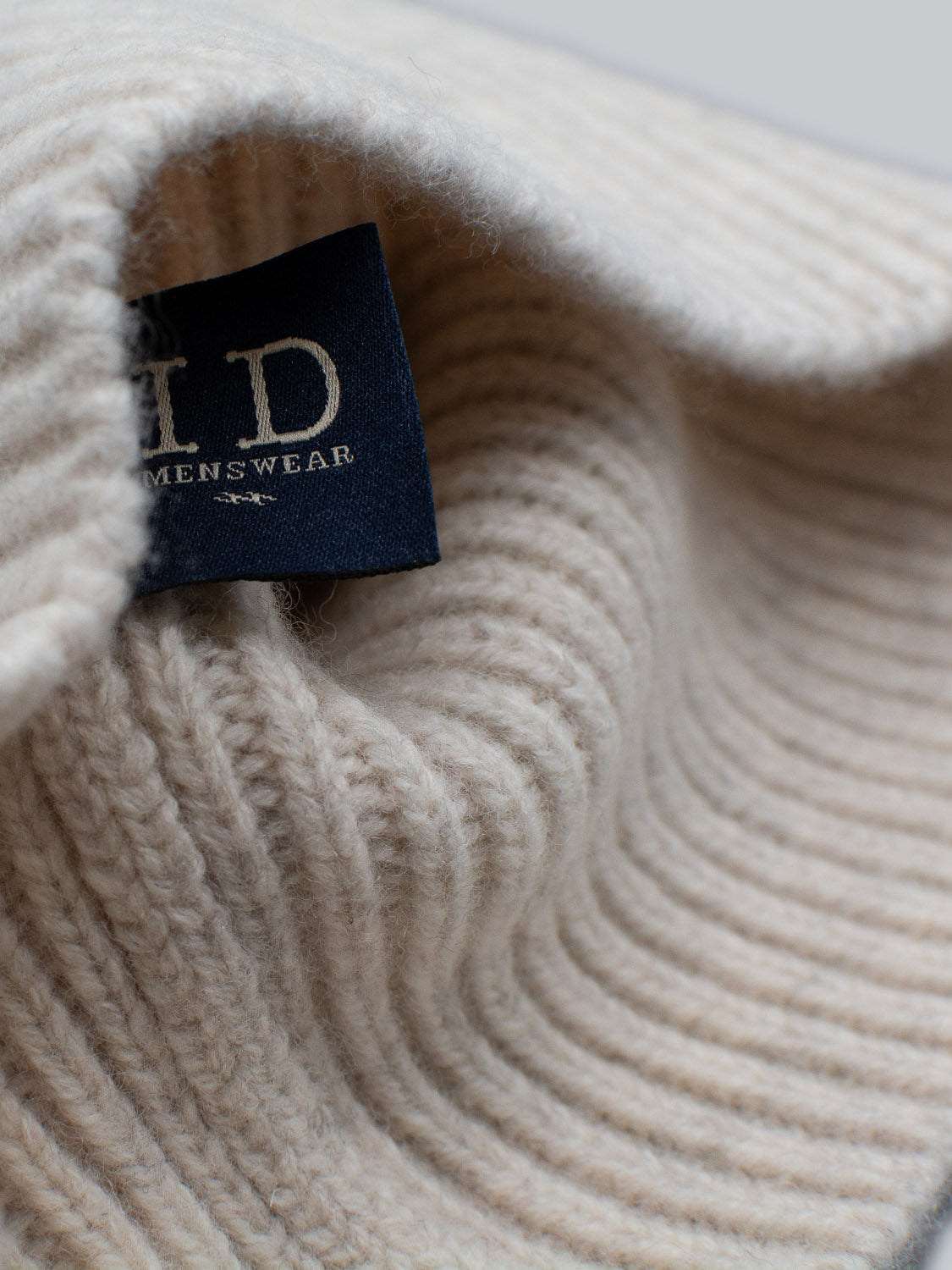 A close-up of a cream-colored, 100% wool knitted fabric from the Chamonix Wool Beanie by ID Menswear reveals its soft, ribbed texture with intricate patterns and natural insulation. A blue ID tag is visible amidst the fabric.