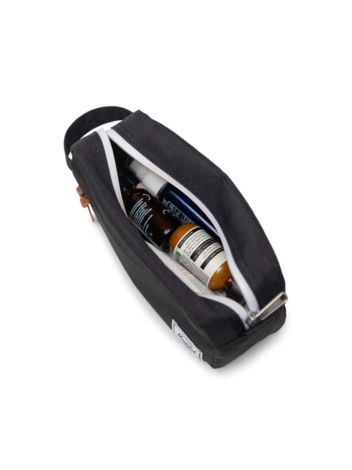 The Herschel Chapter Small Travel Kit, in black with a white zipper, is partially open to reveal bottles and personal care items, serving as a sleek organizer and shown against a white background.