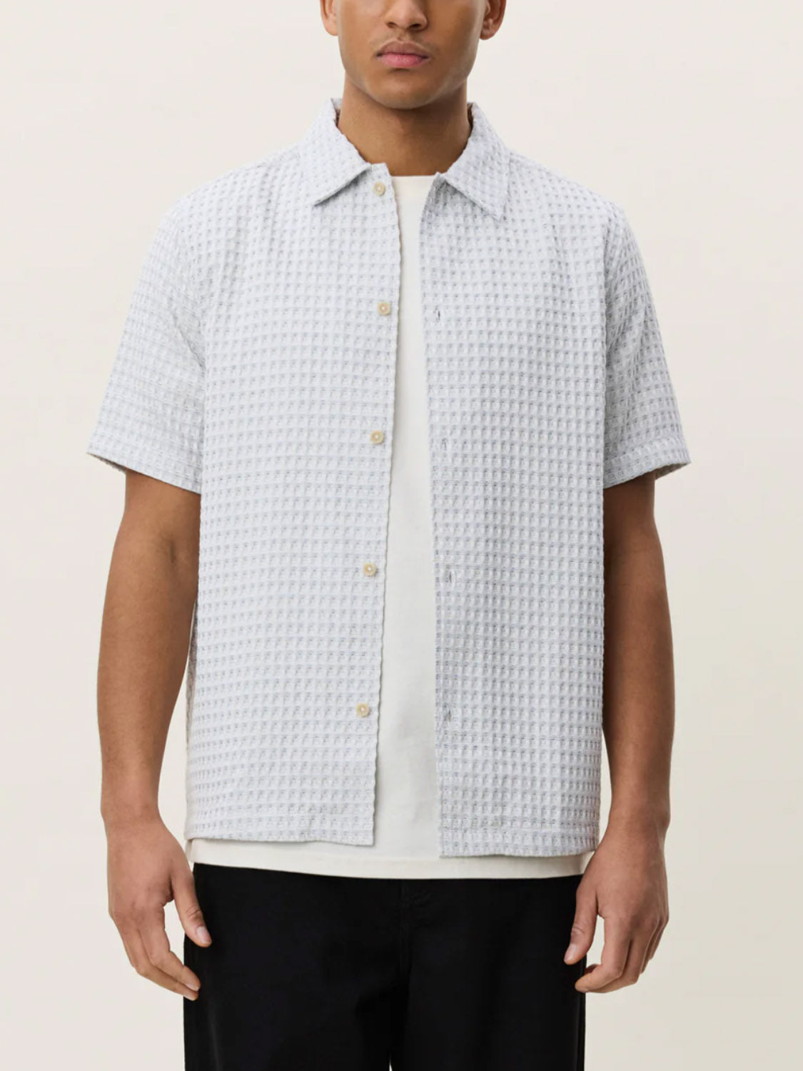 A person is wearing the Les Deux Check Seersucker SS Shirt in light grey, styled over a white t-shirt and black pants, standing against a plain light background.