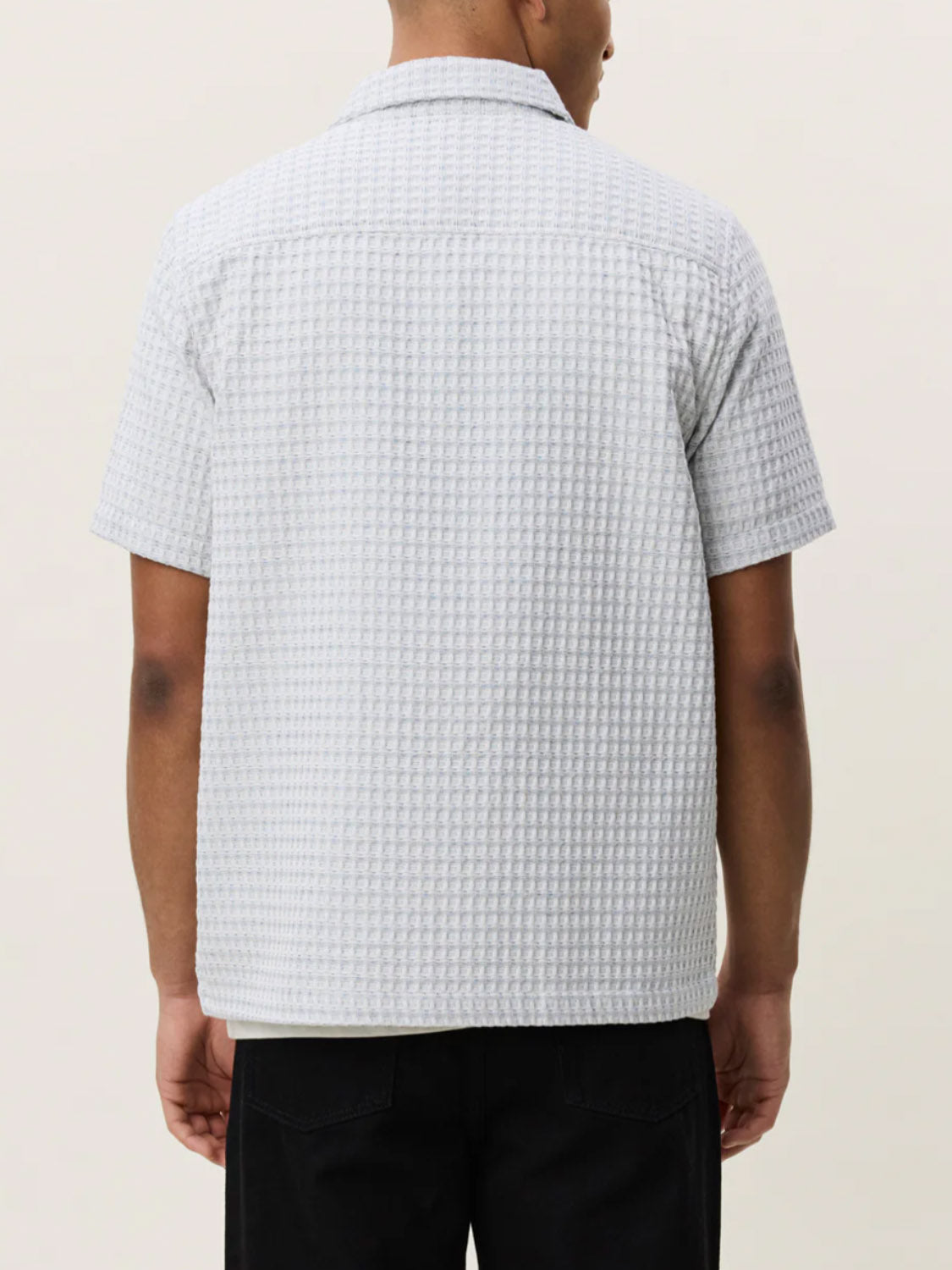 A person is shown from the back against a plain, light-colored background wearing Les Deux's Check Seersucker SS Shirt in light gray with short sleeves and a loose fit, paired with black pants.