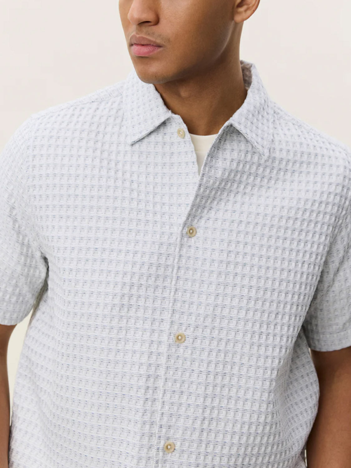 A man wears the Les Deux Check Seersucker SS Shirt, a light grey, loose-fit, waffle-textured button-up over a white undershirt. His face is partially visible against a plain, neutral background.