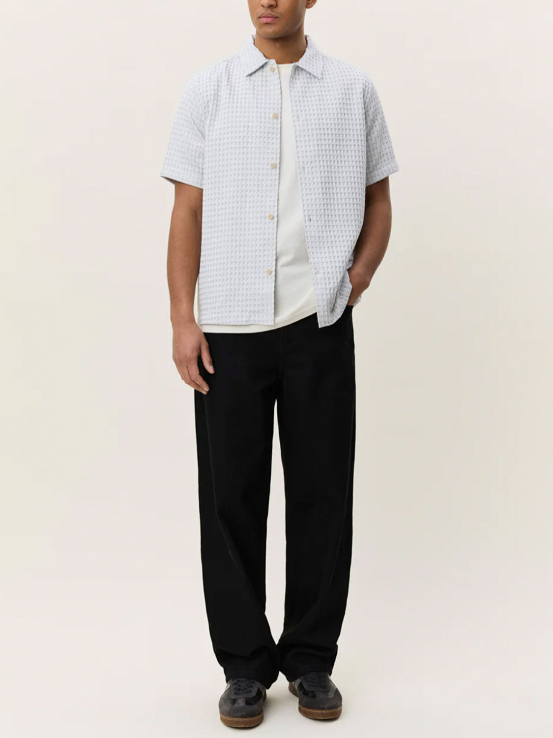 Wearing the Les Deux Check Seersucker SS Shirt over a white T-shirt with loose black pants and black shoes, a person stands against a plain light background.