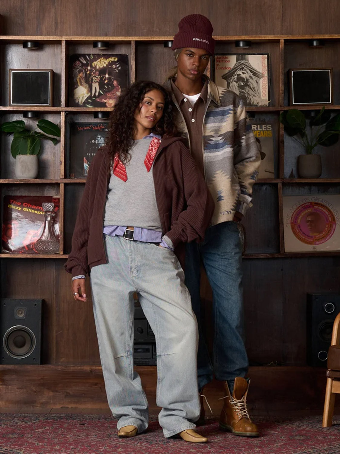 Two people stand by a wood-paneled wall with vinyl records and plants. One wears baggy jeans, a sweater, and scarf; the other sports jeans, boots, and a patterned jacket over the Kuwalla Chenille Polo with a beanie.