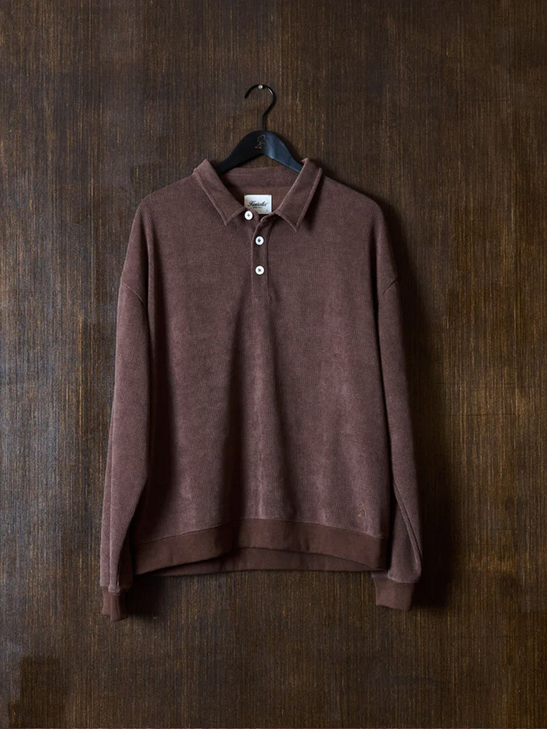 The Kuwalla Chenille Polo, a brown relaxed long-sleeve pullover with three white buttons, hangs on a black hanger against a dark brown textured wall.