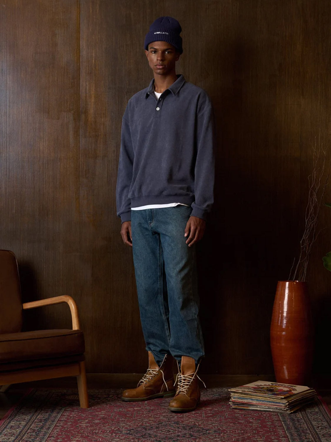 Indoors on a patterned rug, a person wears the Kuwalla Chenille Polo with blue jeans, a navy beanie, and tan lace-up boots. Behind them: a wooden chair, stacked magazines, and a tall brown vase with twigs.