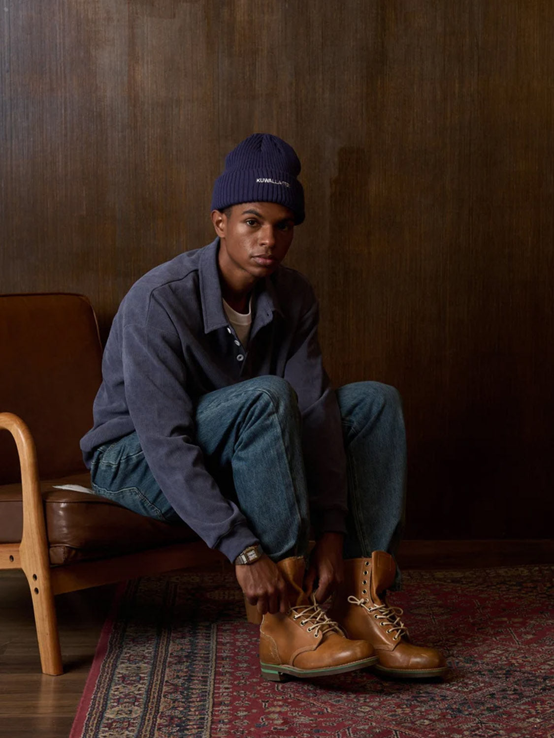 A man wears the Kuwalla Chenille Polo, navy beanie, jeans, and tan boots, sitting on a chair atop a patterned rug with a wooden wall behind him as he looks at the camera and ties his laces.