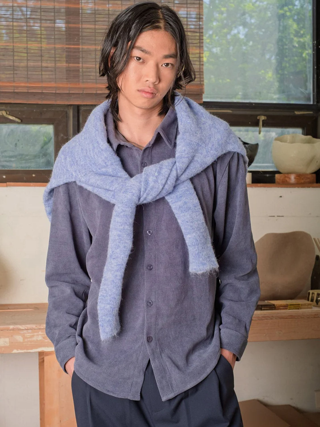 A young person with long dark hair stands indoors, hands in pockets, wearing the Kuwalla Chenille Shirt with a light blue sweater draped over their shoulders. Windows and pottery-filled shelves are visible in the background.