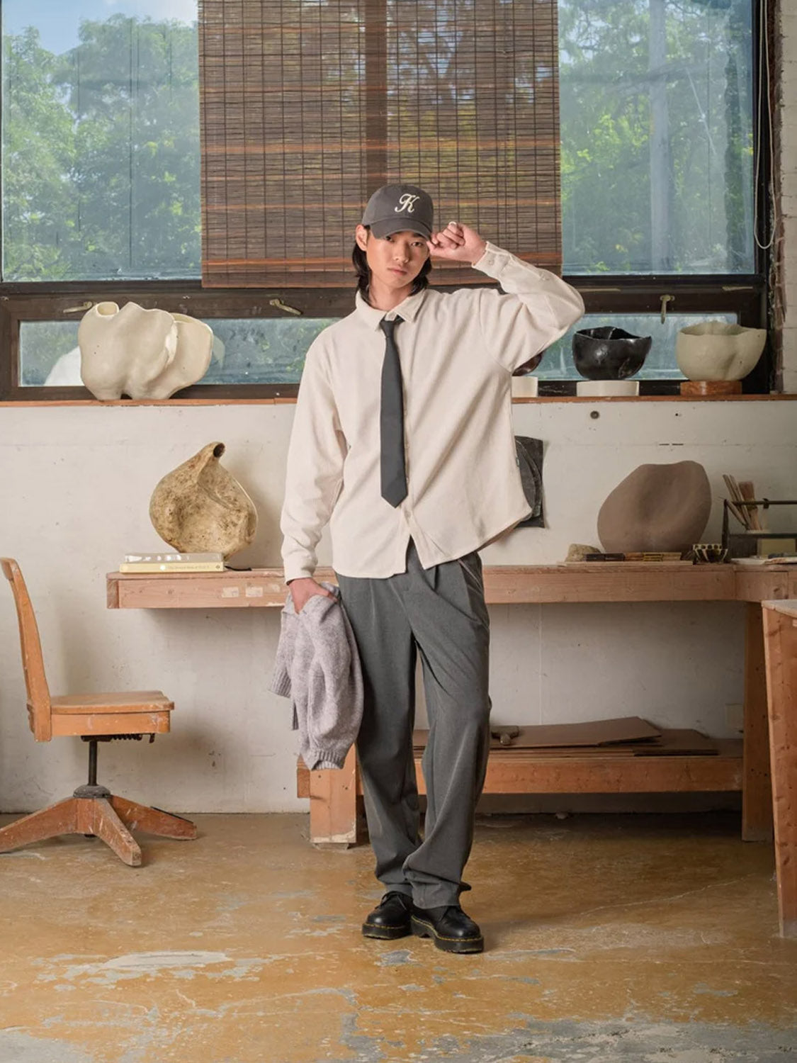 A person in an art studio wears a Kuwalla Chenille Shirt in beige, a gray cap, black tie, gray pants, and holds a gray sweater. Sculptures, art materials, and wooden furniture appear in the background.