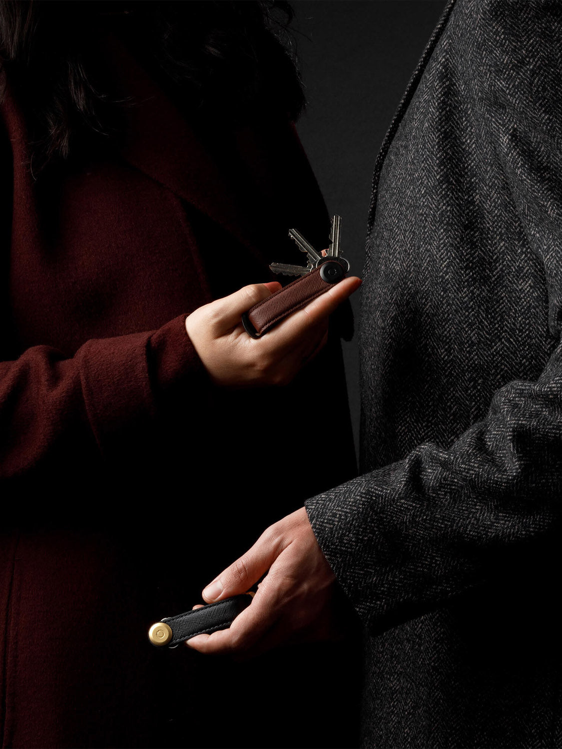 Two people in dark coats stand close together, one holding the Orbitkeys Key Organizer Saffiano Leather and the other with a compact cylindrical Saffiano leather accessory. Both are shown from the shoulders down against a black background.