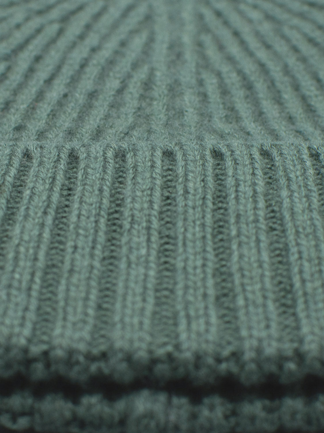 Close-up of green ribbed knit fabric from the Cho Oyu Merino Wool Fishermans Beanie by ID Menswear, featuring vertical textured lines. The soft, fuzzy material promises warmth and comfort typical of merino wool, making it a perfect winter accessory with the cozy feel of classic knitwear.