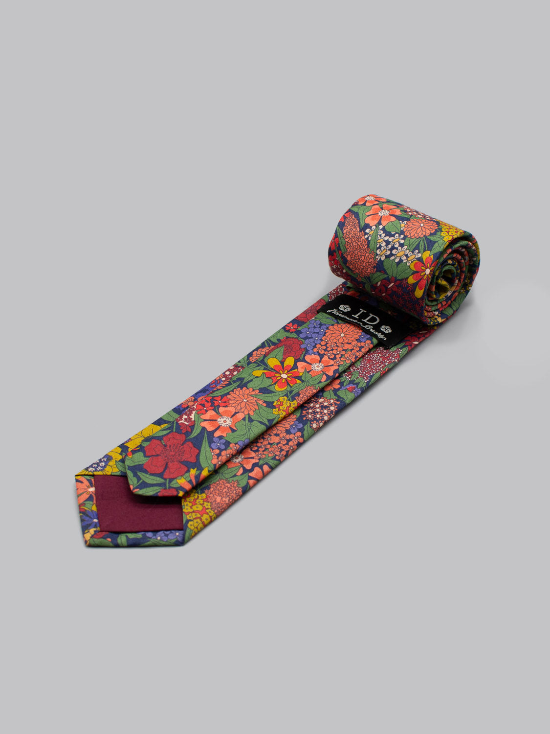 The Liberty of London Tana Lawn™ Ciara Tie by ID Menswear features a vibrant floral pattern in red, yellow, orange, pink, and green on a dark background; handmade in Brooklyn and shown rolled up on a light gray surface.