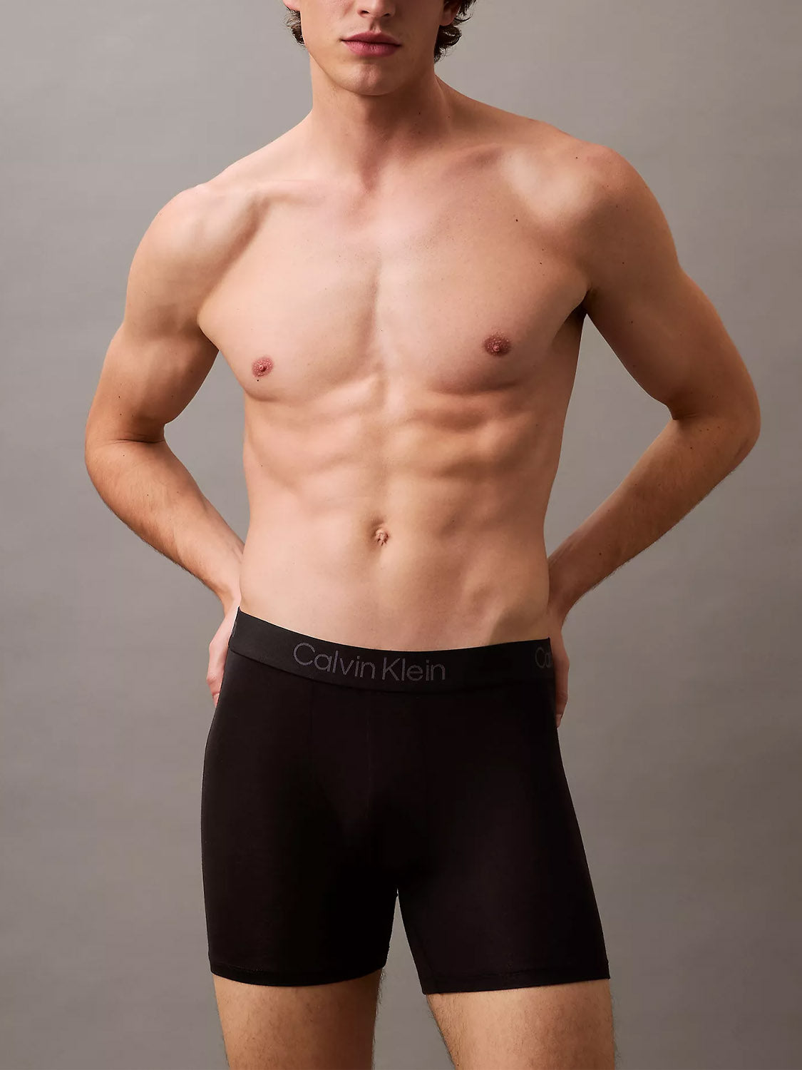 A shirtless man stands against a plain gray background, wearing Calvin Klein NB4193 Ultra Soft Modal 3-Pack Boxer Briefs in black, with his hands on his hips. His face is partially out of frame.