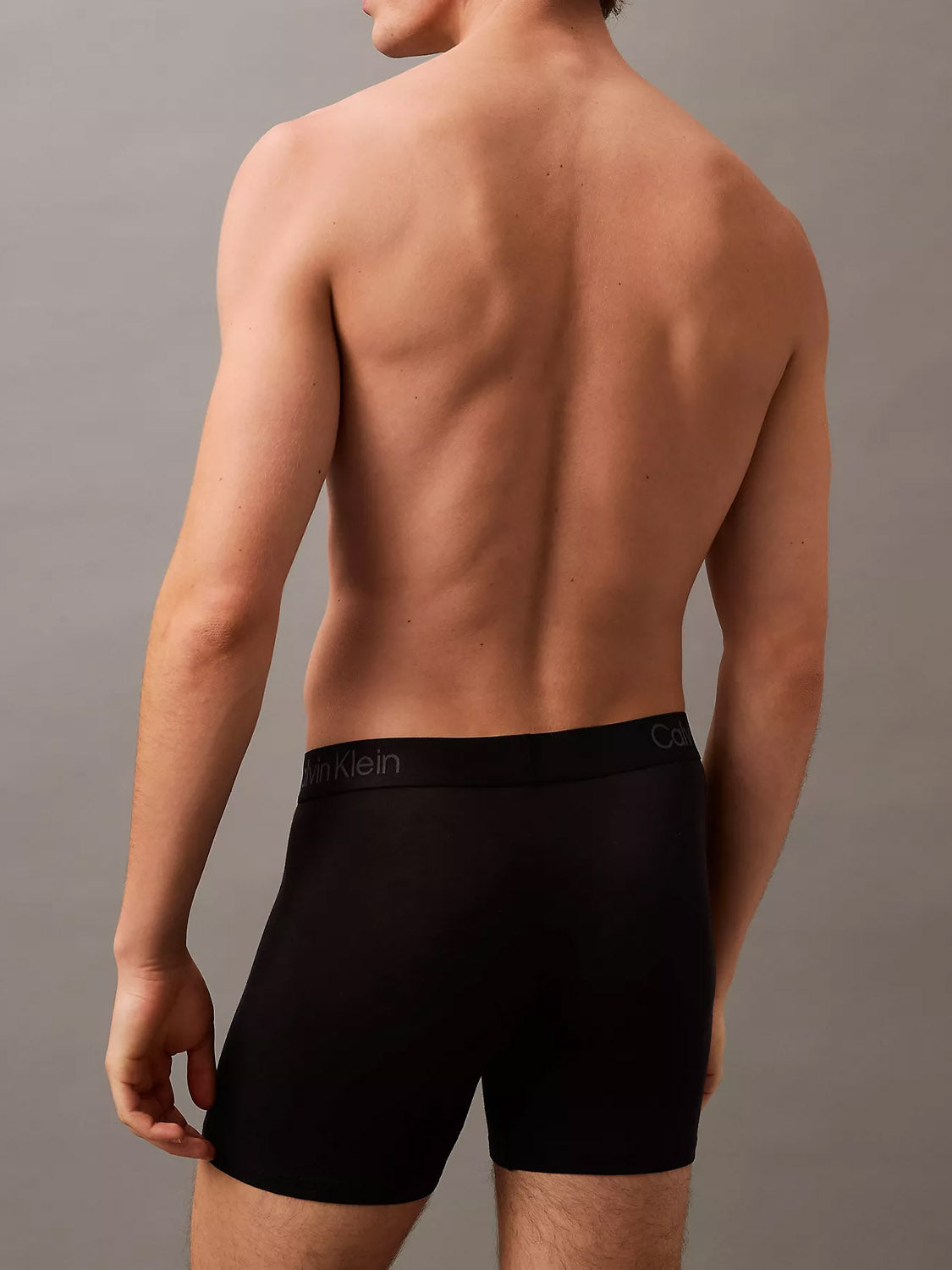 A shirtless person faces away from the camera, modeling Calvin Klein’s NB4193 Ultra Soft Modal 3-Pack Boxer Briefs against a plain, neutral background.