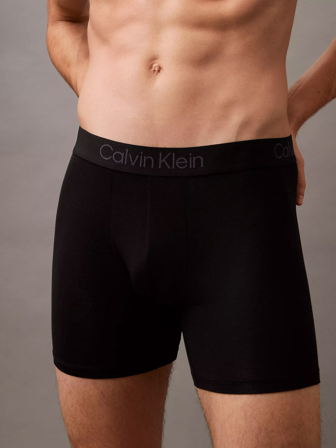 A model showcases the Calvin Klein NB4193 Ultra Soft Modal 3-Pack Boxer Brief, standing with hands on hips against a plain background. The focus is on the torso and upper thighs, emphasizing the comfort and breathability of this underwear.