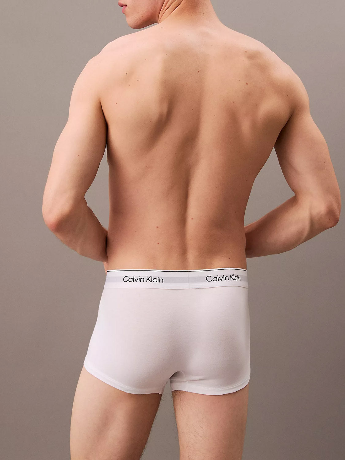 A shirtless man faces away from the camera, wearing Calvin Klein NB4389 Icon Cotton Stretch 3-Pack Low Rise Trunk boxer briefs, featuring a stitch-free waistband, set against a plain, light-colored background.