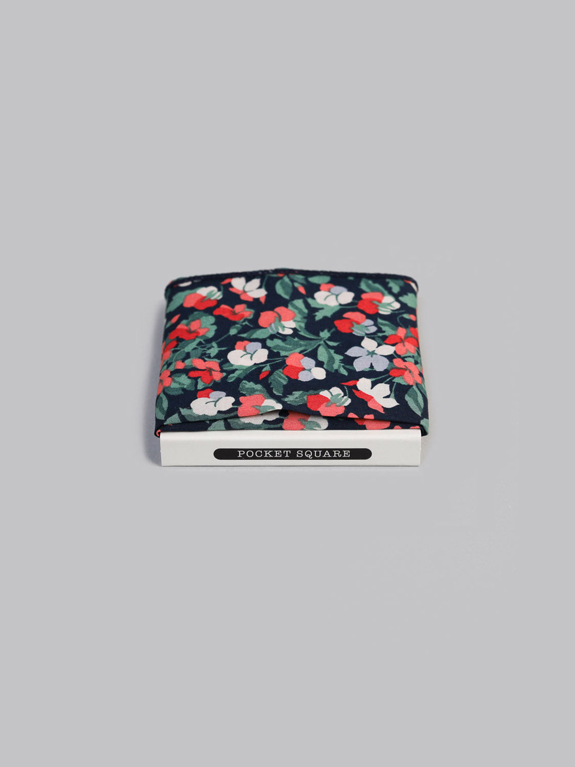 A Liberty™ Classic Pocket Square by ID Menswear, crafted from red, white, and green floral Tana Lawn cotton, is expertly handmade in Brooklyn. It rests folded in a white POCKET SQUARE box against a gray background.
