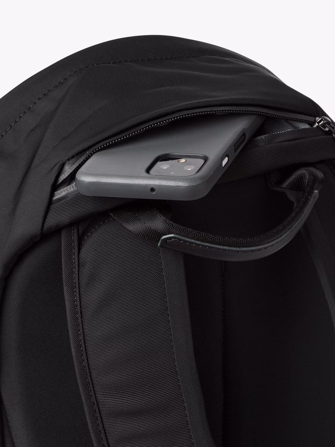 A close-up of the Bellroy Classic Compact Backpack - 16L shows its zippered pocket partially open with a black smartphone tucked inside.