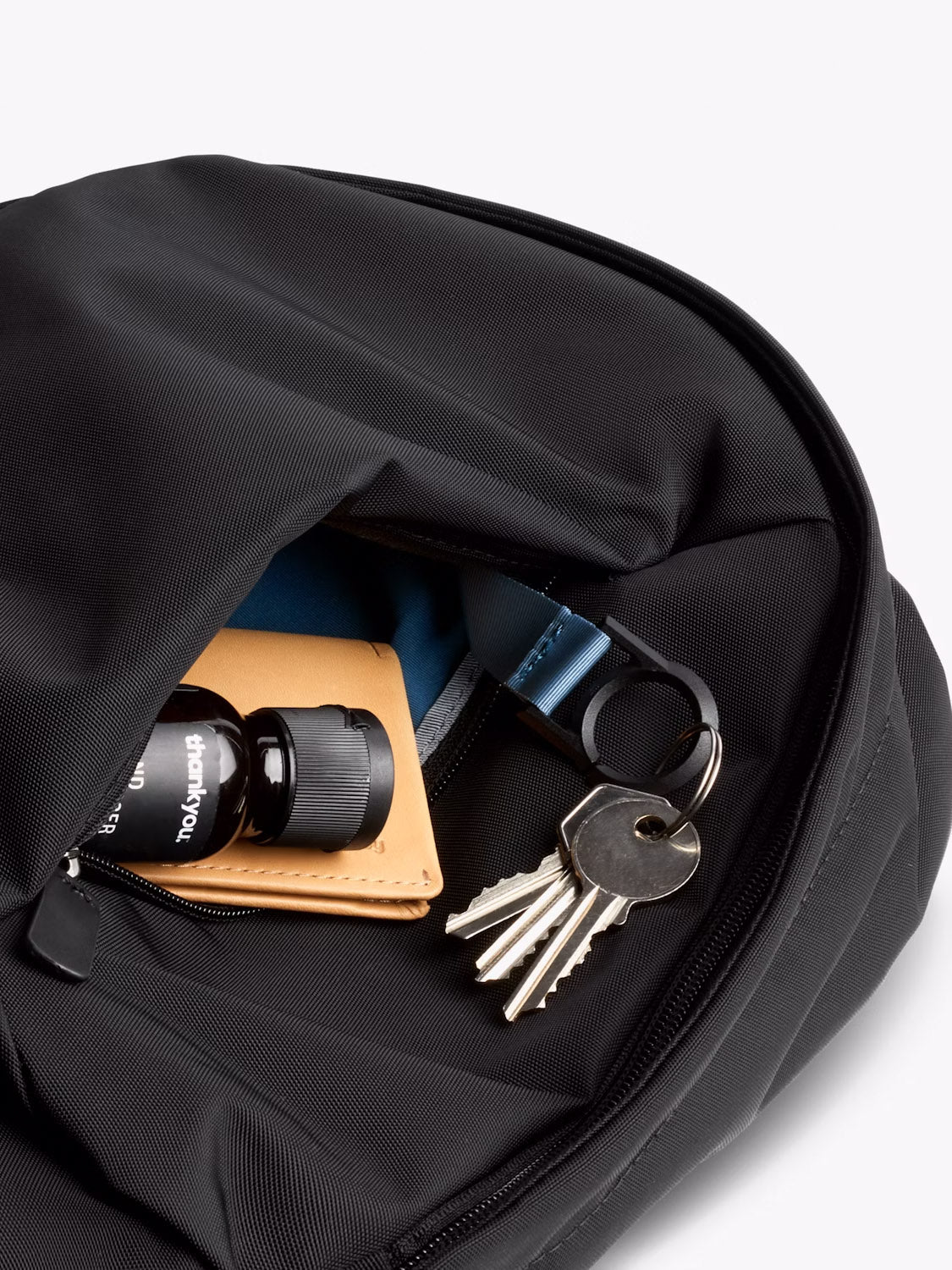 A close-up of the Bellroy Classic Compact Backpack - 16L, showing its unzipped pocket with a tan wallet, small black bottle, and a silver keyring with three keys. The partially visible interior highlights the bag’s organized design.