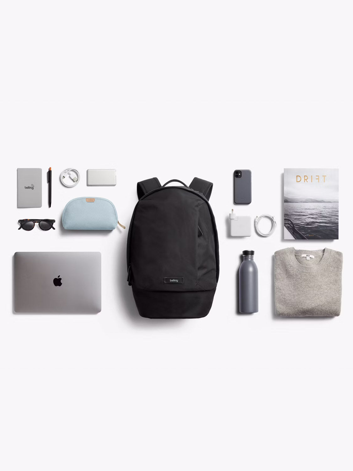 A flat lay of travel essentials featuring Bellroy’s Classic Compact Backpack - 16L in black, alongside a laptop, sweater, water bottle, sunglasses, notebook, pen, pouch, phone, charging cable, adapter, wallet and Drift magazine.