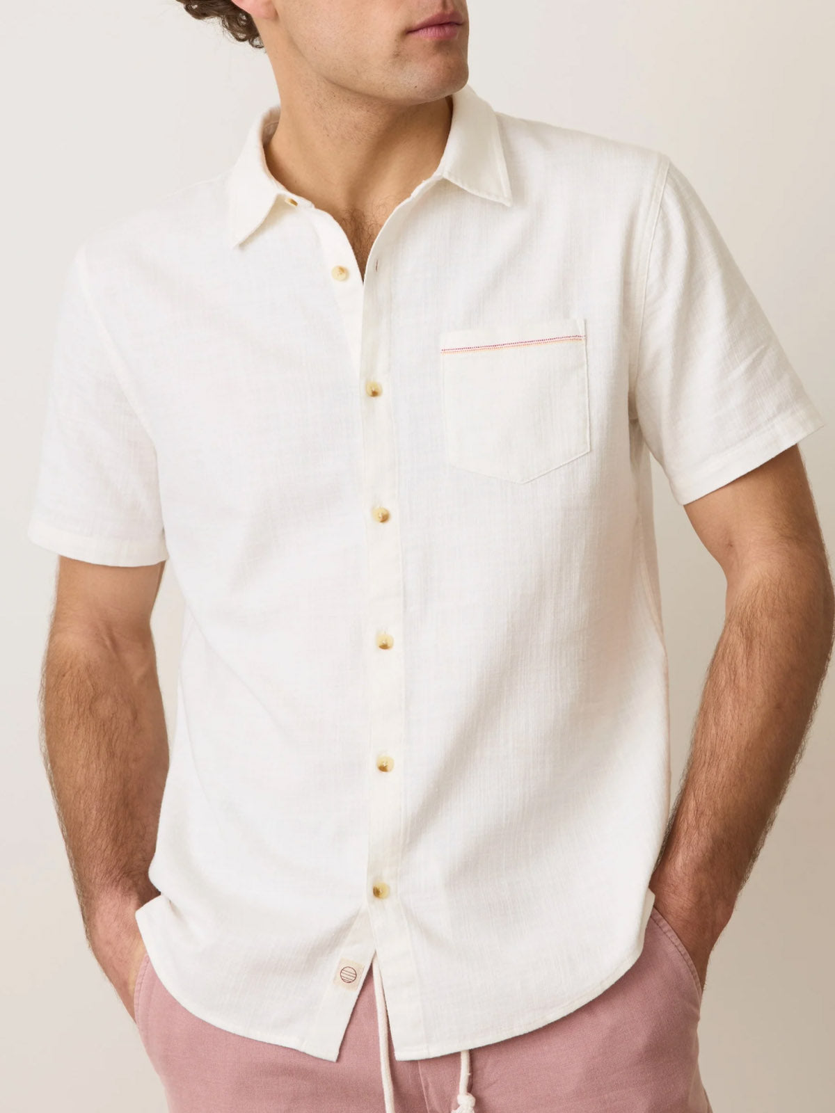 A man wears a Marine Layer Classic Stretch Selvage Shirt (short-sleeve, front pocket) with light pink shorts, standing with one hand in his pocket; his face is partially obscured.