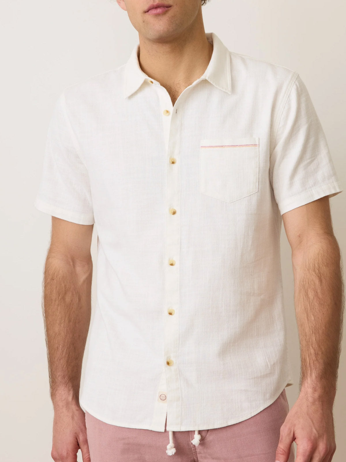 A man wearing a Marine Layer Classic Stretch Selvage Shirt in off-white with a front pocket and light buttons, paired with light pink pants, stands against a plain background. His face is partially out of frame.