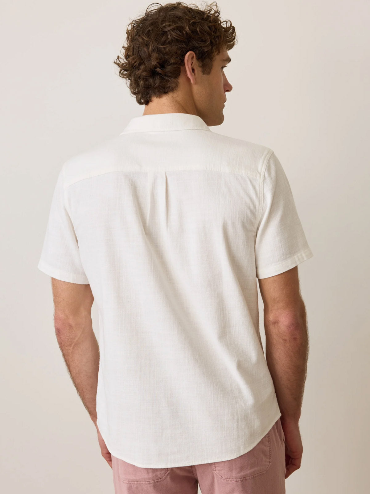 A man with curly brown hair is shown from behind in a Marine Layer Classic Stretch Selvage Shirt with short sleeves and light pink pants, standing against a plain light background.