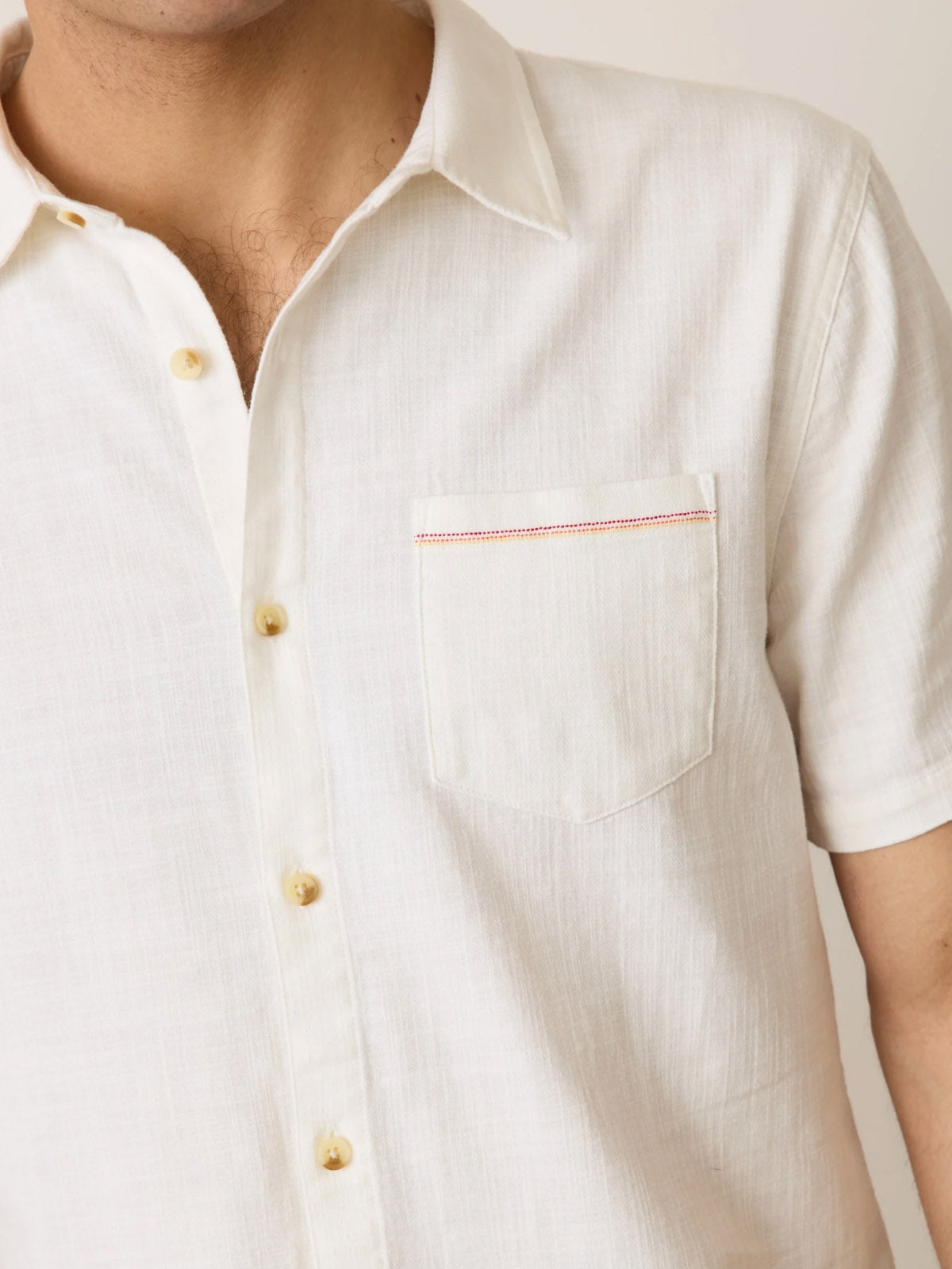 A close-up of a person wearing the Marine Layer Classic Stretch Selvage Shirt: an off-white, short-sleeve shirt with a classic fit, chest pocket, and red stitched accent. Only part of their face and upper body are shown.