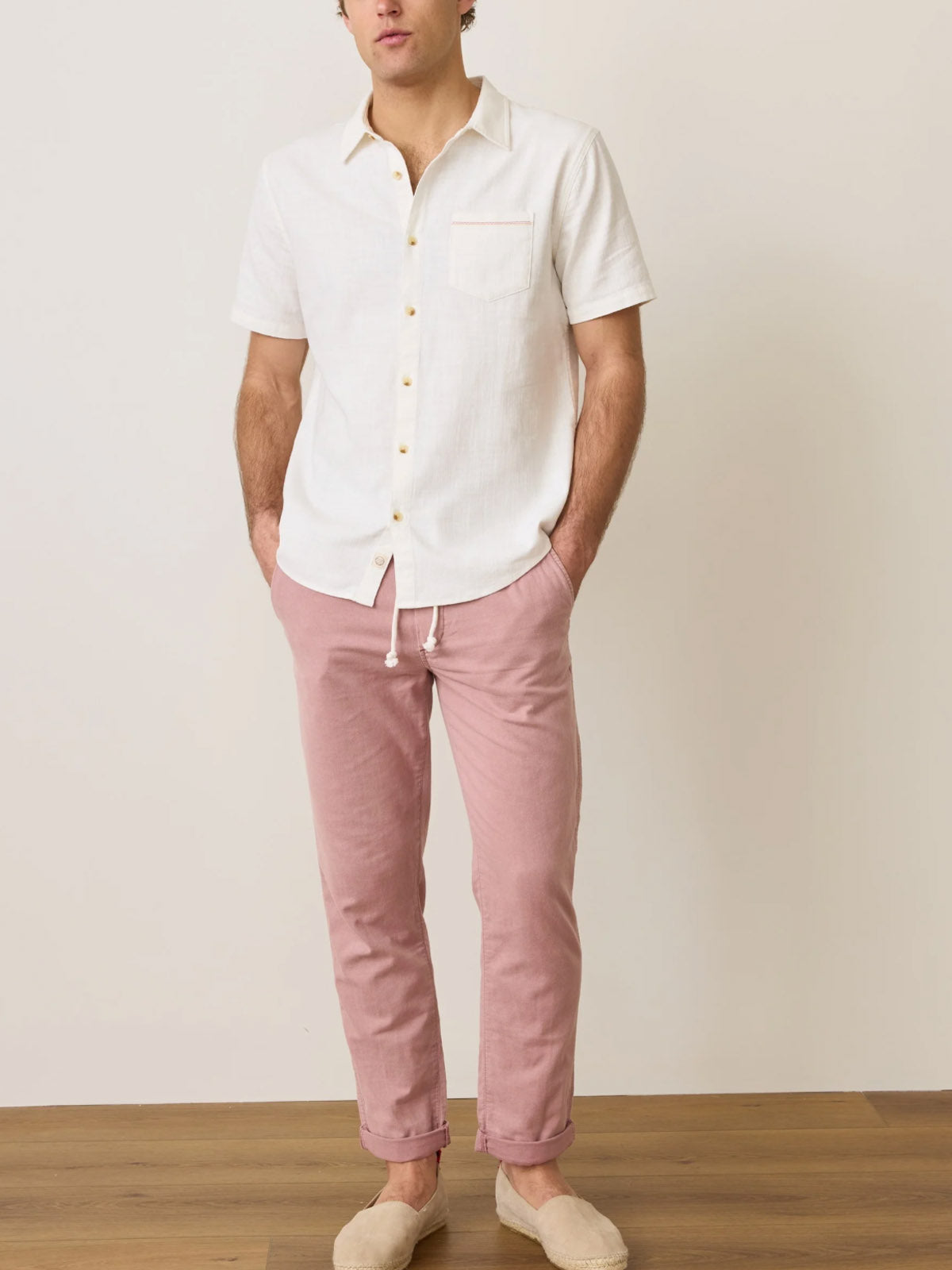 A man stands indoors in a Marine Layer Classic Stretch Selvage Shirt and light pink drawstring pants with beige slip-on shoes, hands in pockets, against a plain light wall on a wood floor.