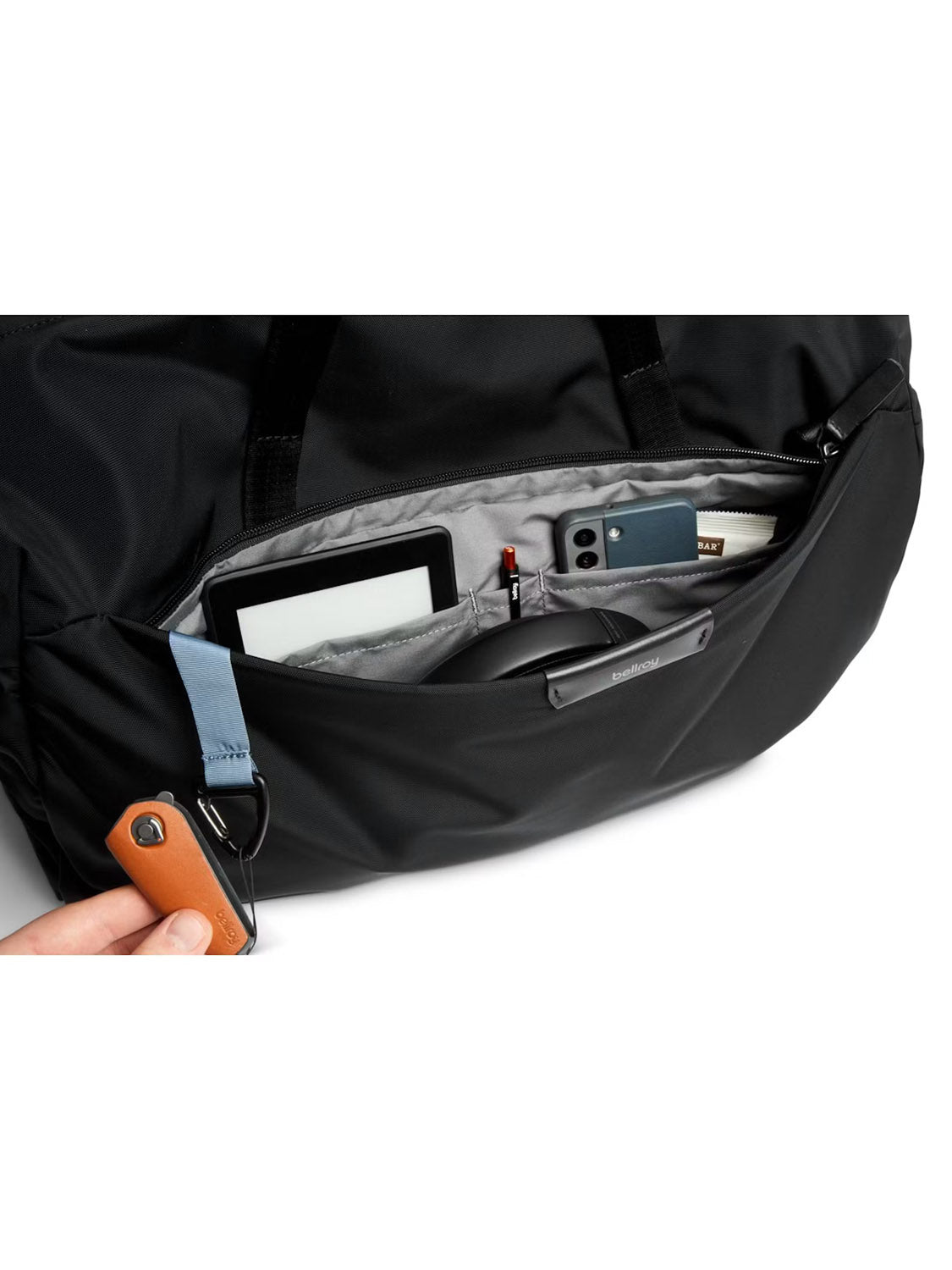 A close-up of an open Bellroy Classic Weekender Duffle Bag - 45L’s front pocket shows organized items—e-reader, smartphone, pen, sunglasses, and wallet. A hand holds a keychain attached to the zipper.