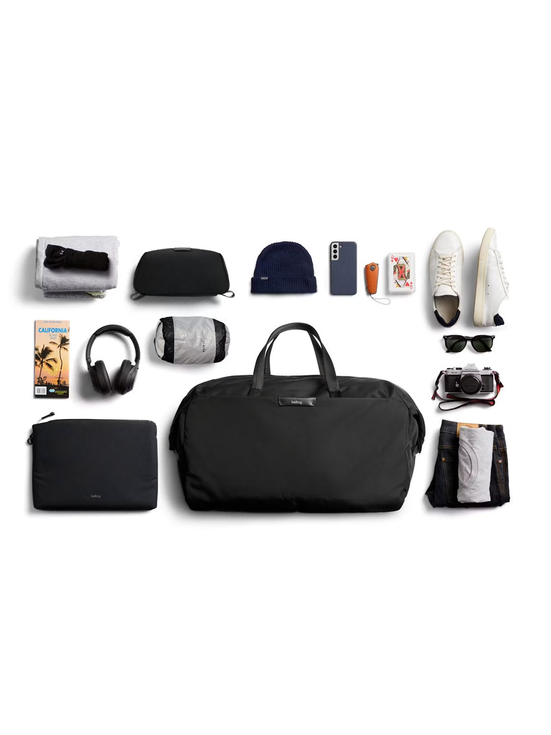 Flat lay of travel essentials featuring the Bellroy Classic Weekender Duffle Bag - 45L with luggage pass-through and laptop sleeve, plus headphones, camera, sneakers, sunglasses, toiletry bag, beanie, wallet, smartphone, playing cards & a travel guide.