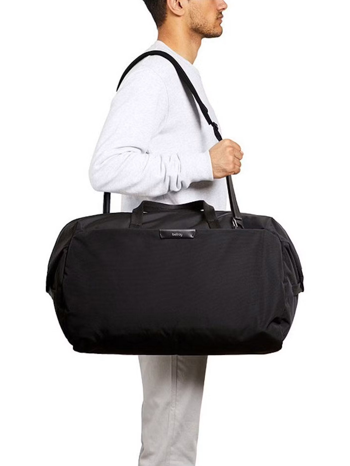A person in a light gray sweater and light pants carries the Bellroy Classic Weekender Duffle Bag - 45L, featuring both a shoulder strap and handles, against a plain white background.