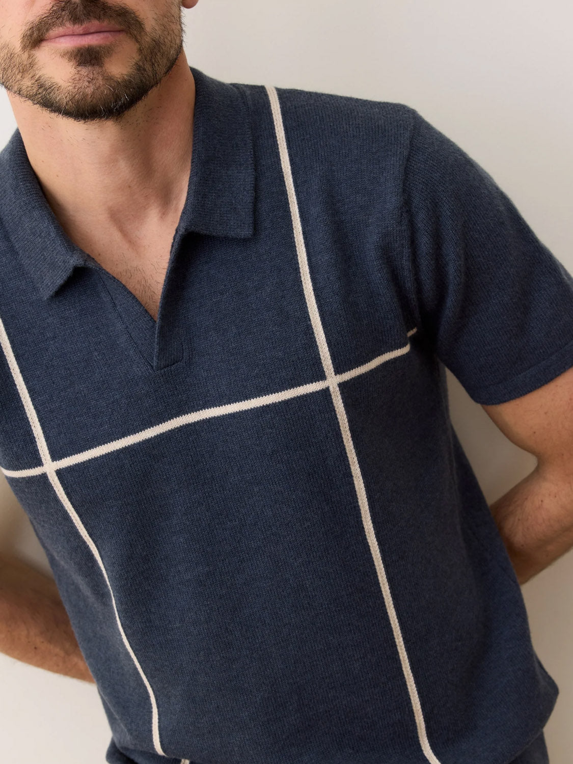 A man with a short beard wears the Marine Layer Cliff Sweater Polo, a navy blue short-sleeve shirt with a collar and white grid lines. He stands before a plain light wall, arms slightly behind him, highlighting contemporary style.