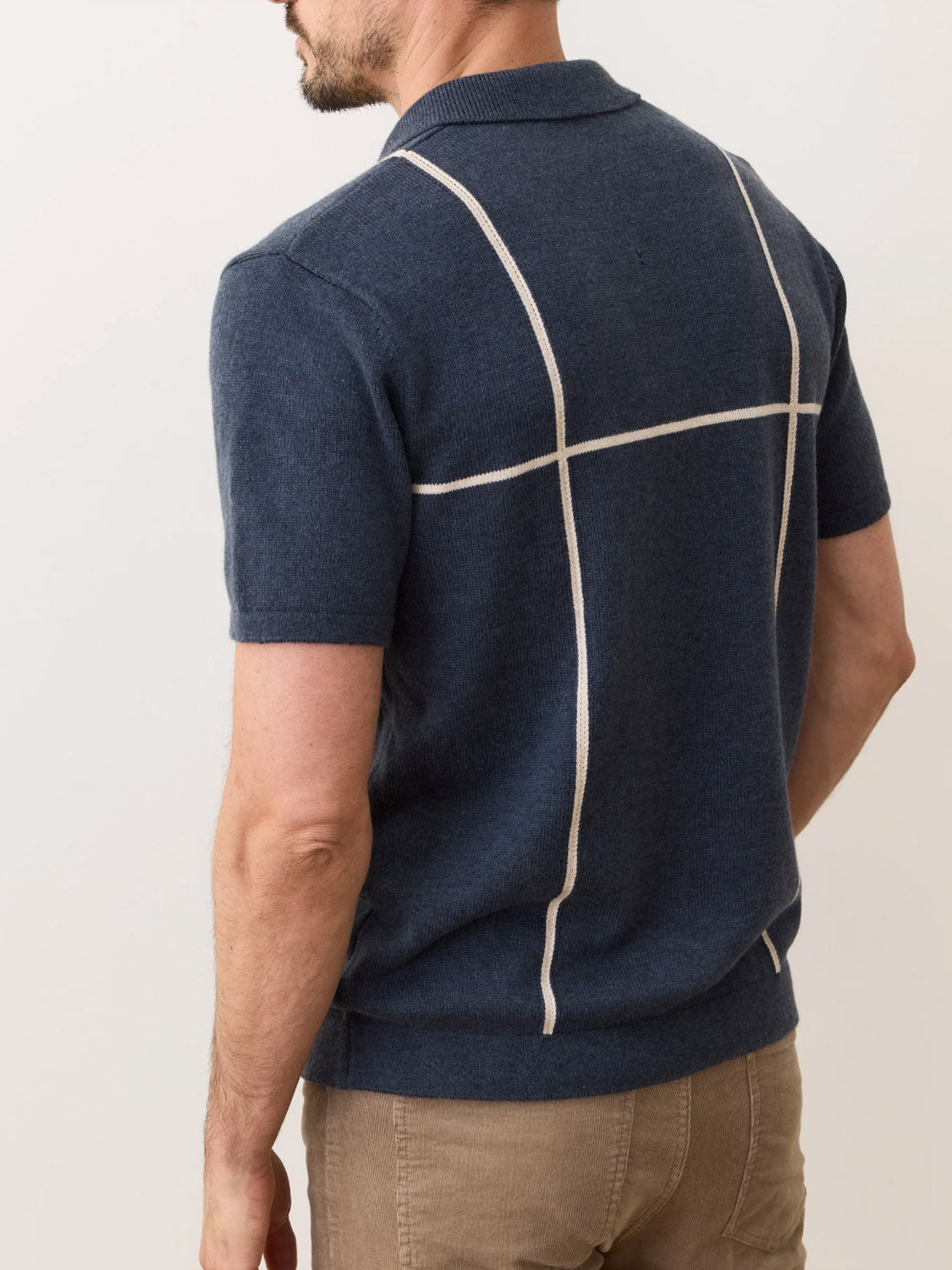 A man is shown from the back wearing the Marine Layer Cliff Sweater Polo, a modern navy blue knit shirt with a collar and thin white grid lines, made from a cotton-nylon-wool blend. He pairs it with light brown pants.