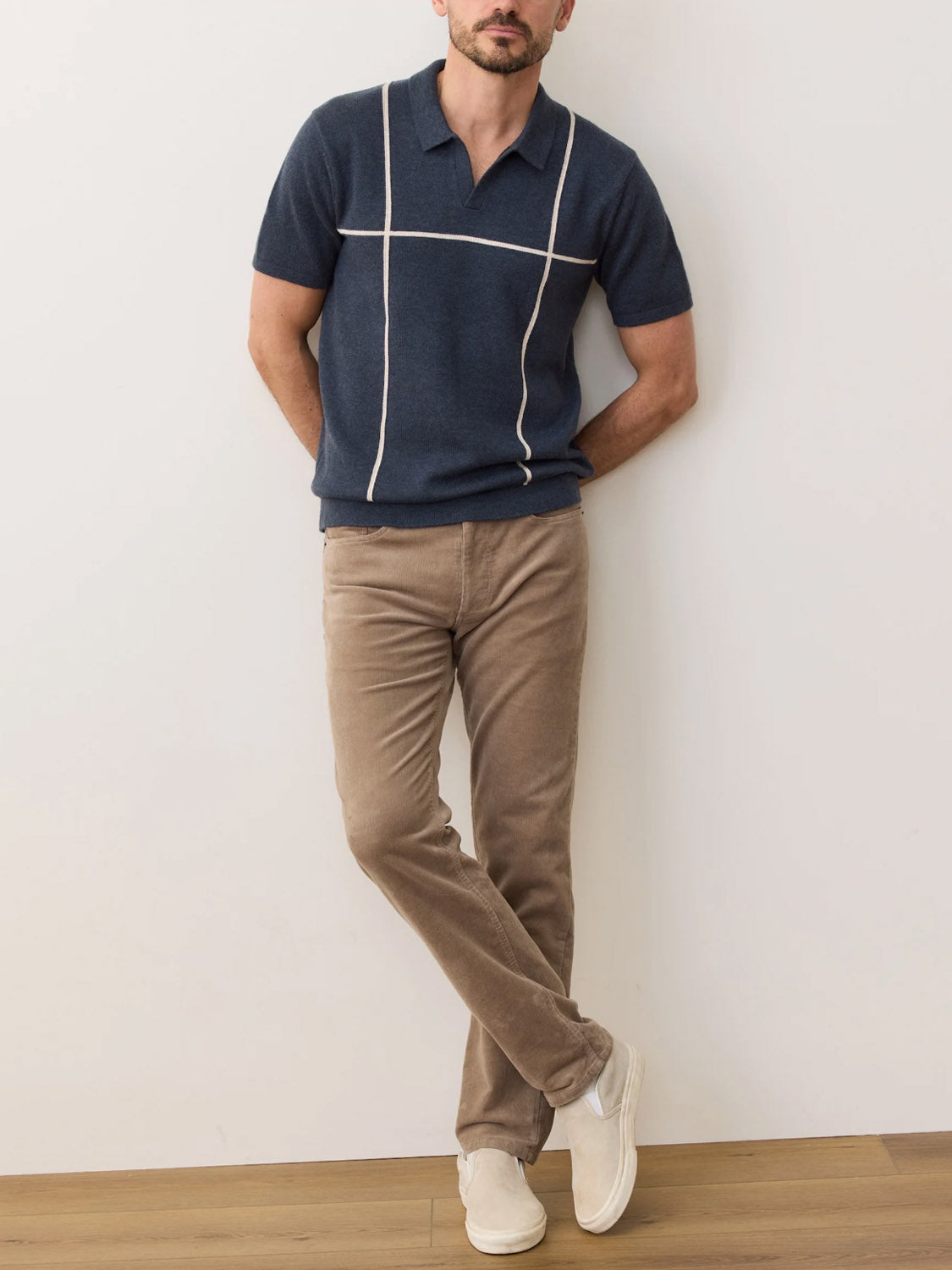 A man stands against a plain wall with arms crossed, wearing the Marine Layer Cliff Sweater Polo in navy with white stripes, tan corduroy pants, and white slip-on shoes, set on a light wood floor.