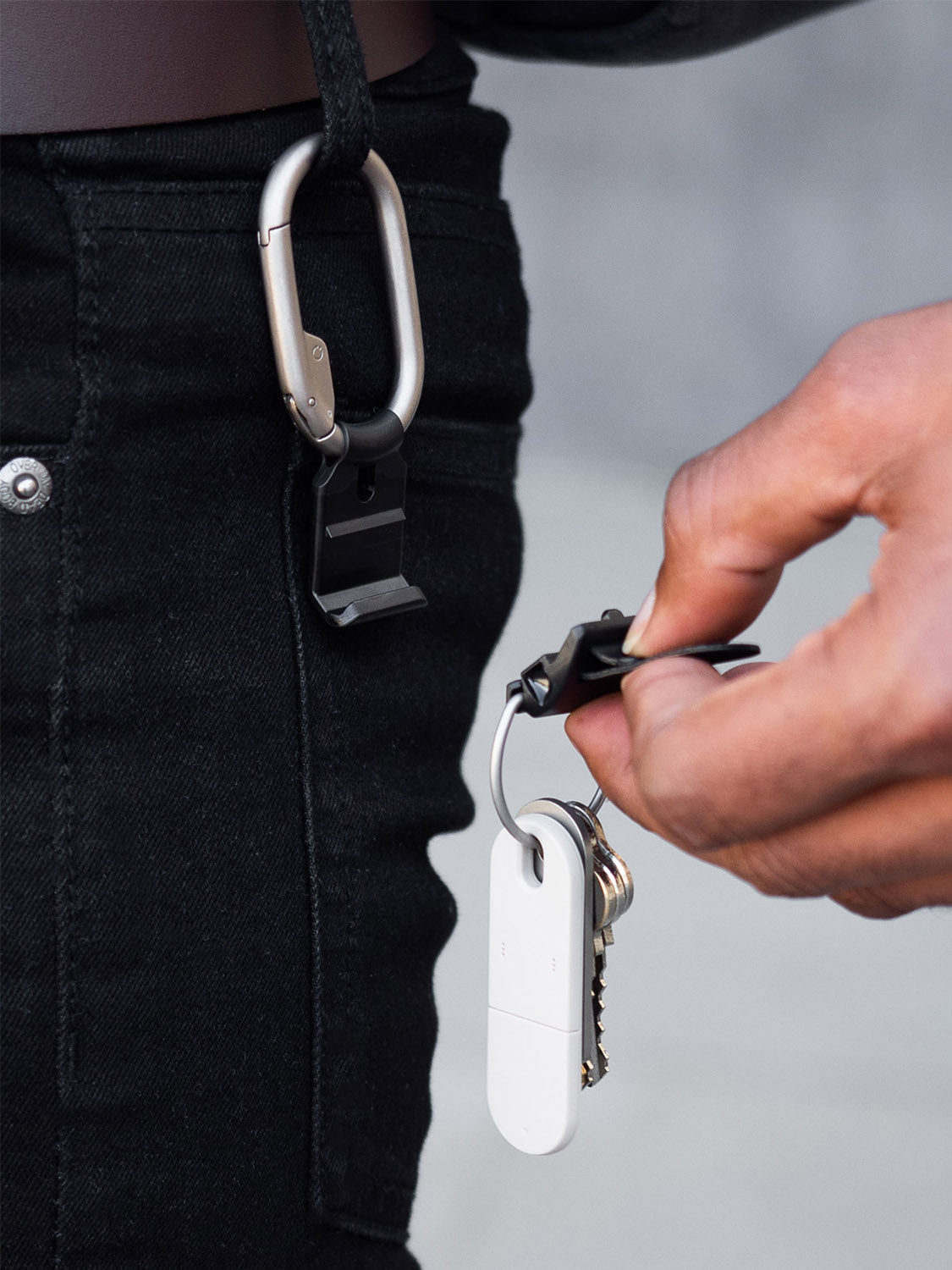 A hand removes the Orbitkeys Clip V2 Keyring with keys and a white fob from a magnetic quick-release belt loop clip attached to black jeans.
