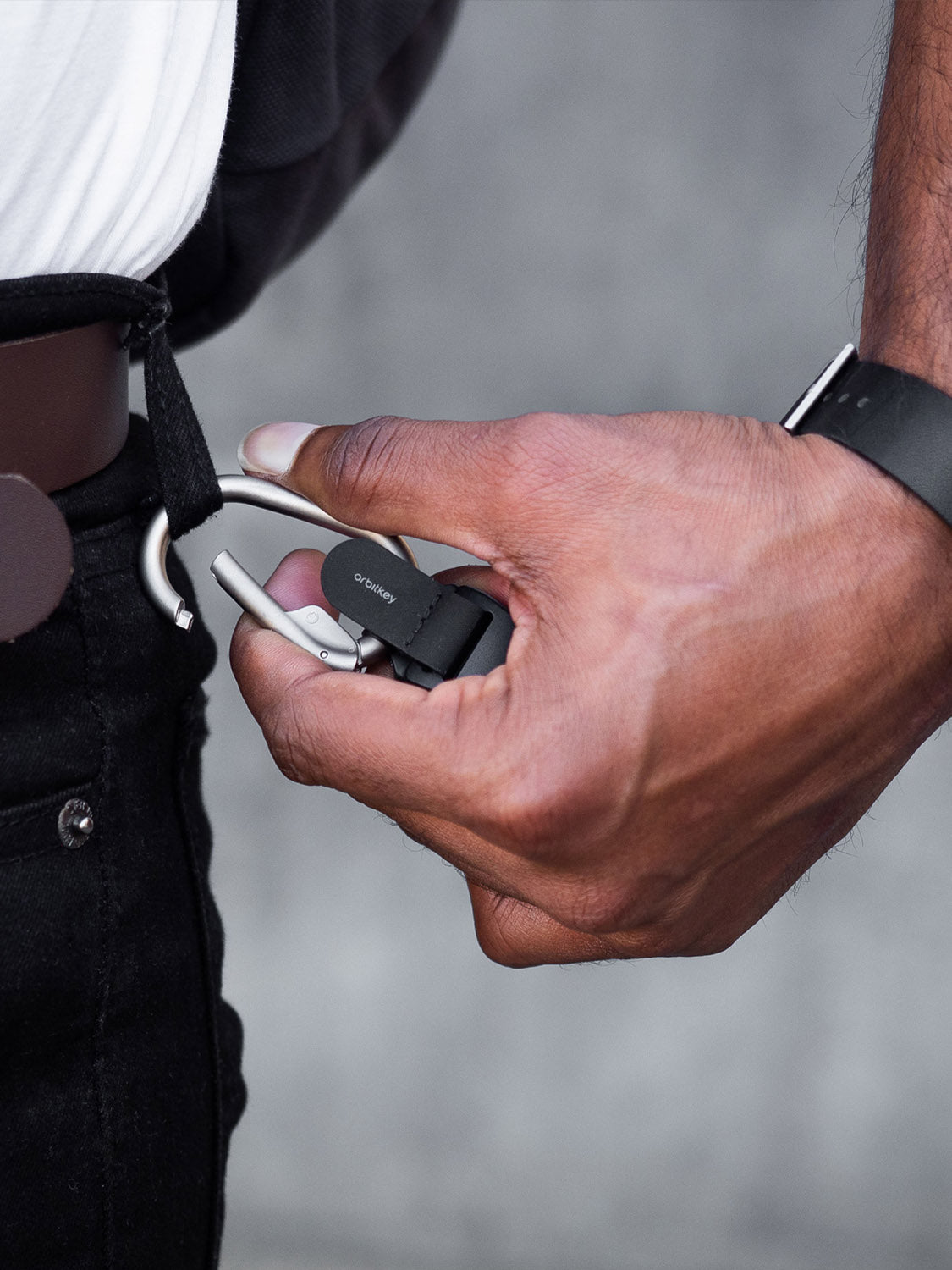 A hand clips the Orbitkeys Clip V2 Keyring with magnetic quick release onto a black jeans belt loop. The person wears a black watch and a white tucked-in shirt. The out-of-focus background is gray.