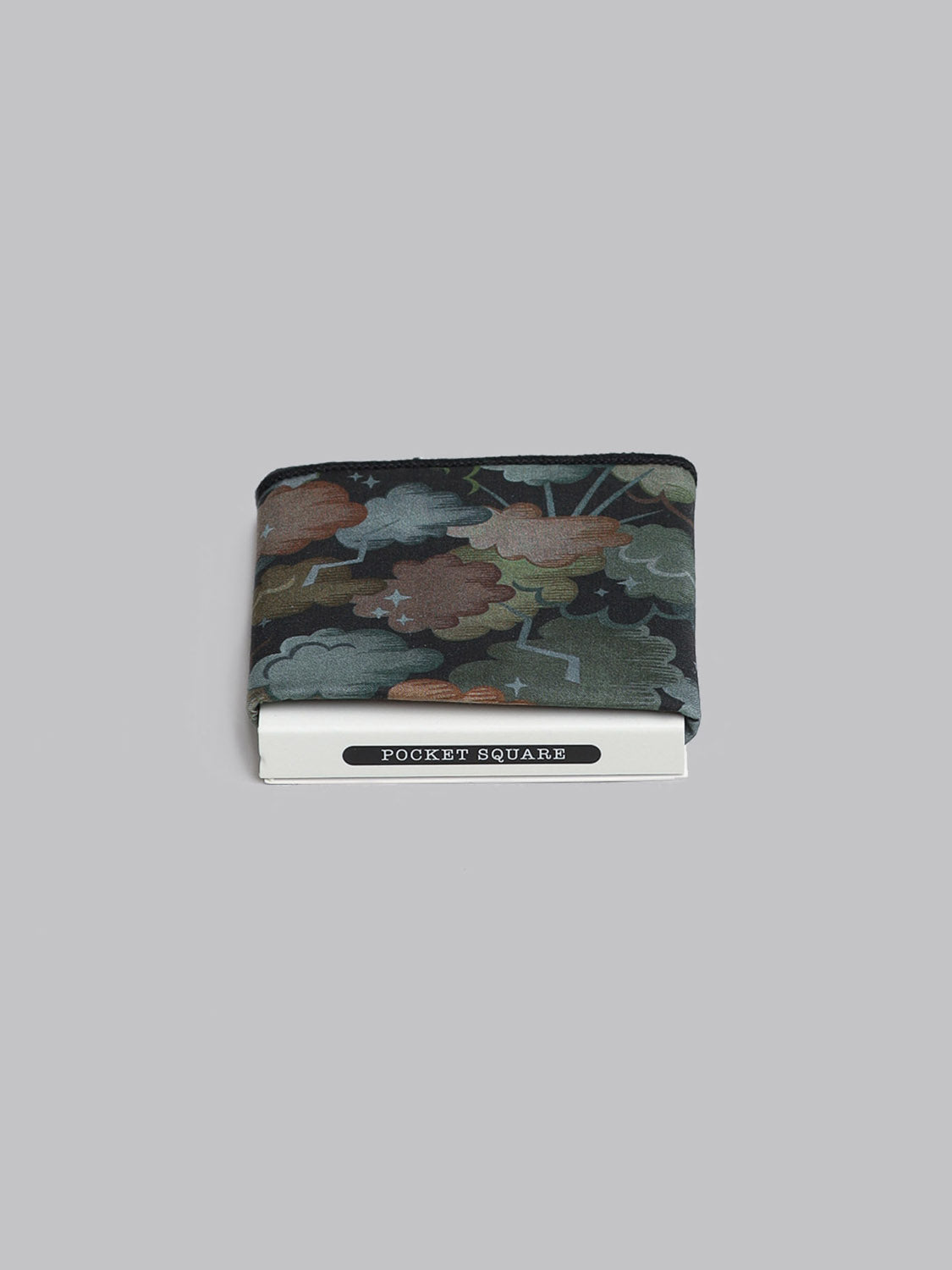 The Liberty™ Cloud Nine Pocket Square by ID Menswear features a dark, multicolored abstract pattern. Handmade in Brooklyn, it is shown folded atop a white POCKET SQUARE box against a plain gray background.