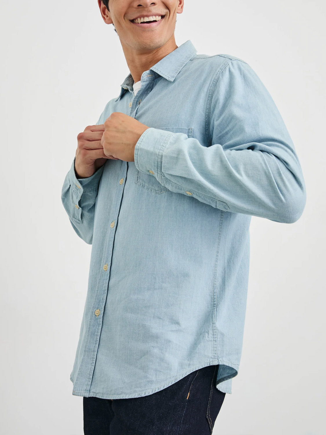 A person smiles as they button up the Rails Colton Dark Wash Indigo Shirt. The focus is on the shirt and their hands, with dark pants partially visible against a light, plain background.