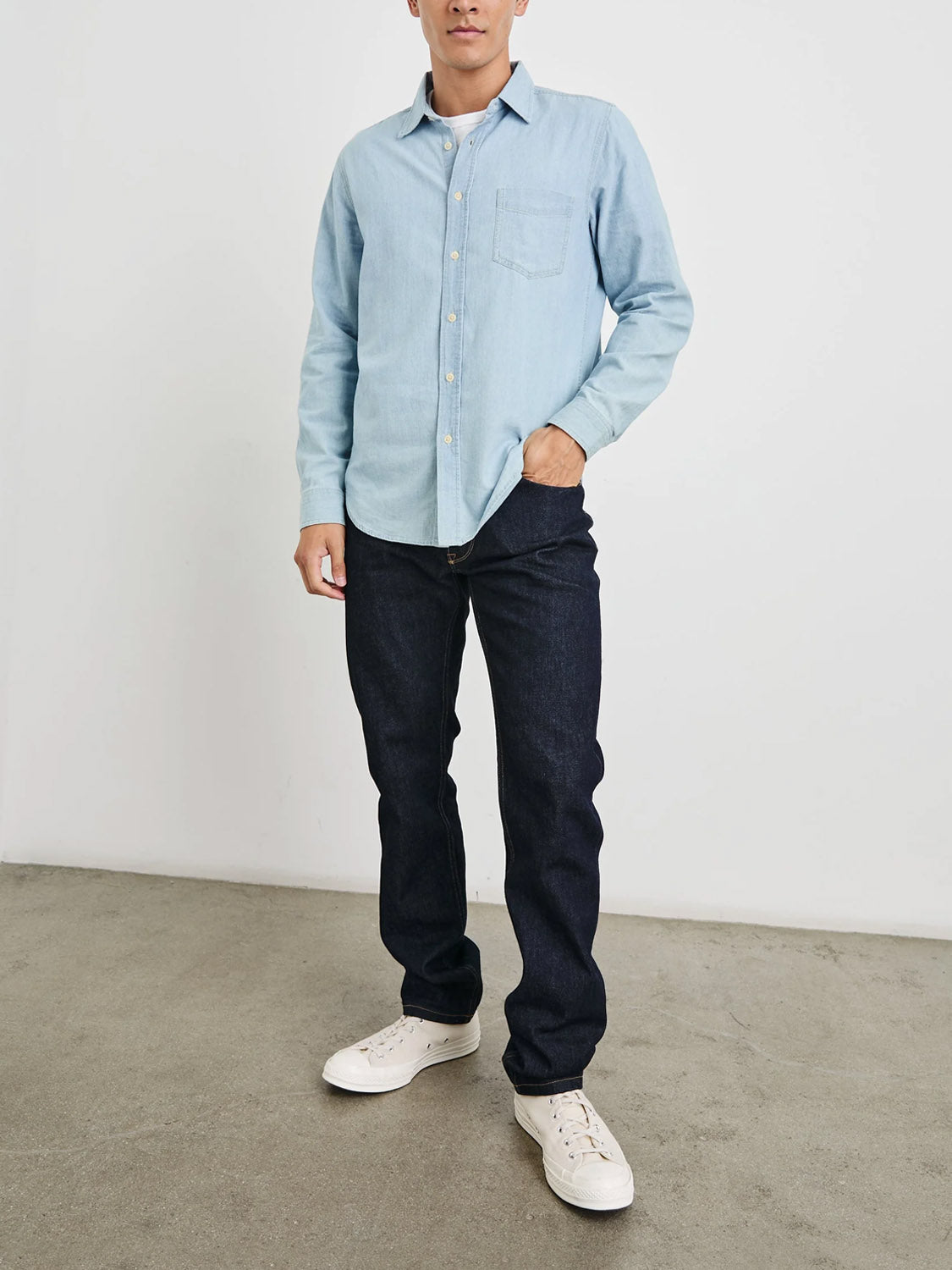 Posing against a plain background, they wear the Rails Colton Dark Wash Indigo Shirt with dark jeans and white sneakers, their right hand casually in their pocket, embodying a vintage-inspired, effortlessly relaxed style.
