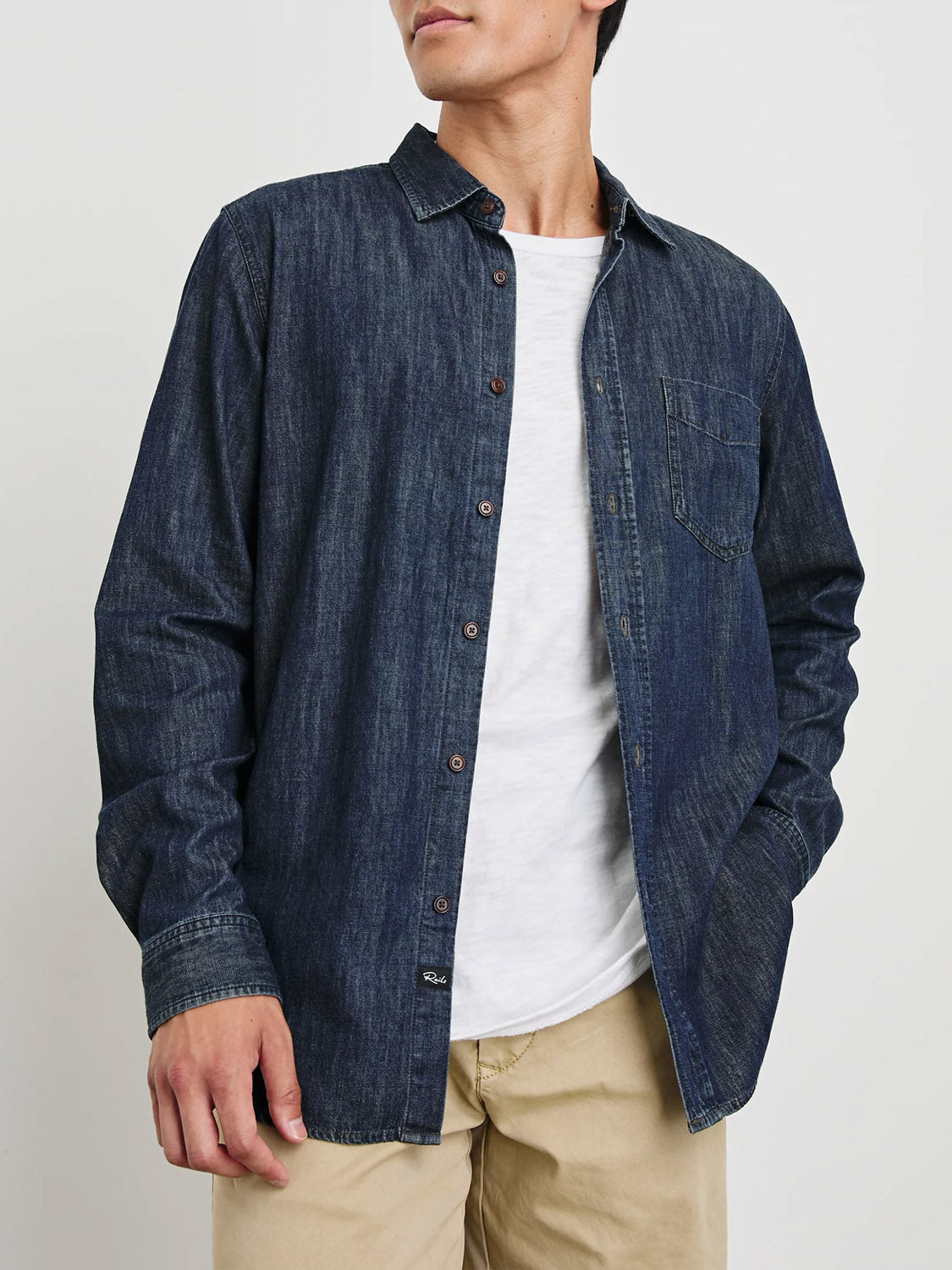 A person wearing a vintage-inspired Rails Colton Dark Wash Indigo Shirt over a white T-shirt and beige pants stands against a plain background. The shirt features a front pocket and brown buttons, while their left hand casually rests in their pants pocket.