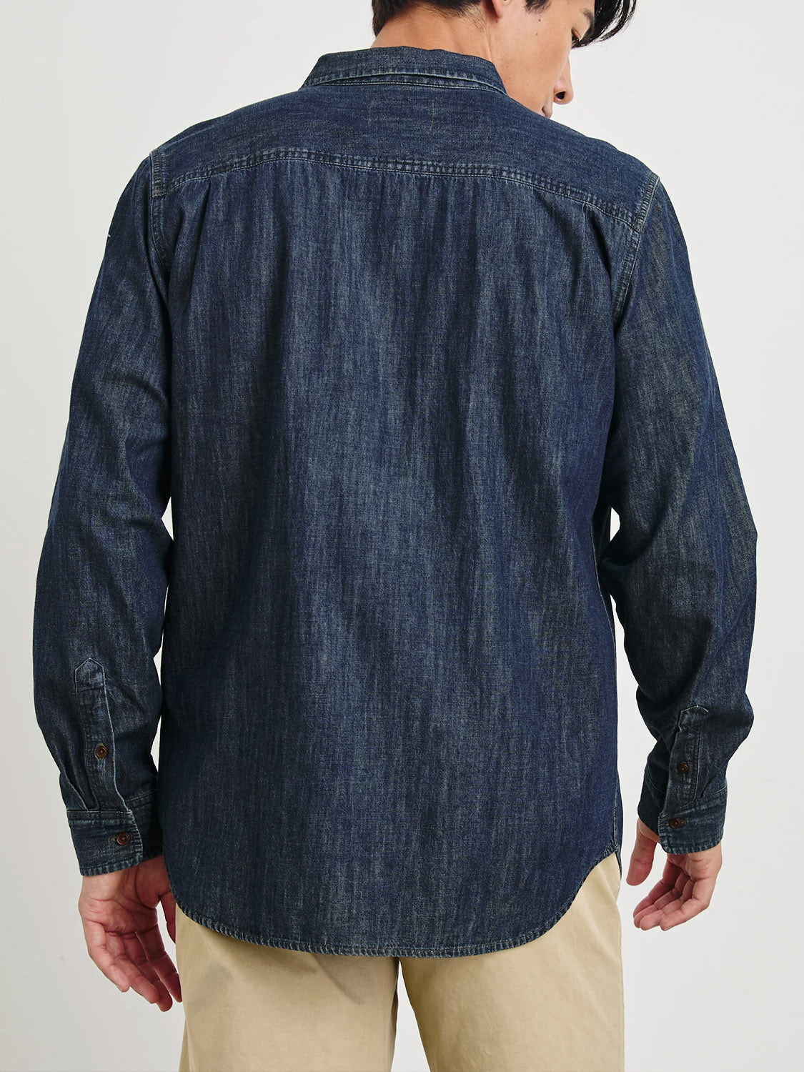A man in a Rails Colton Dark Wash Indigo Shirt, exuding a vintage charm with its casual style and slightly curved hem, pairs it with light khaki pants, standing with his back to the camera.