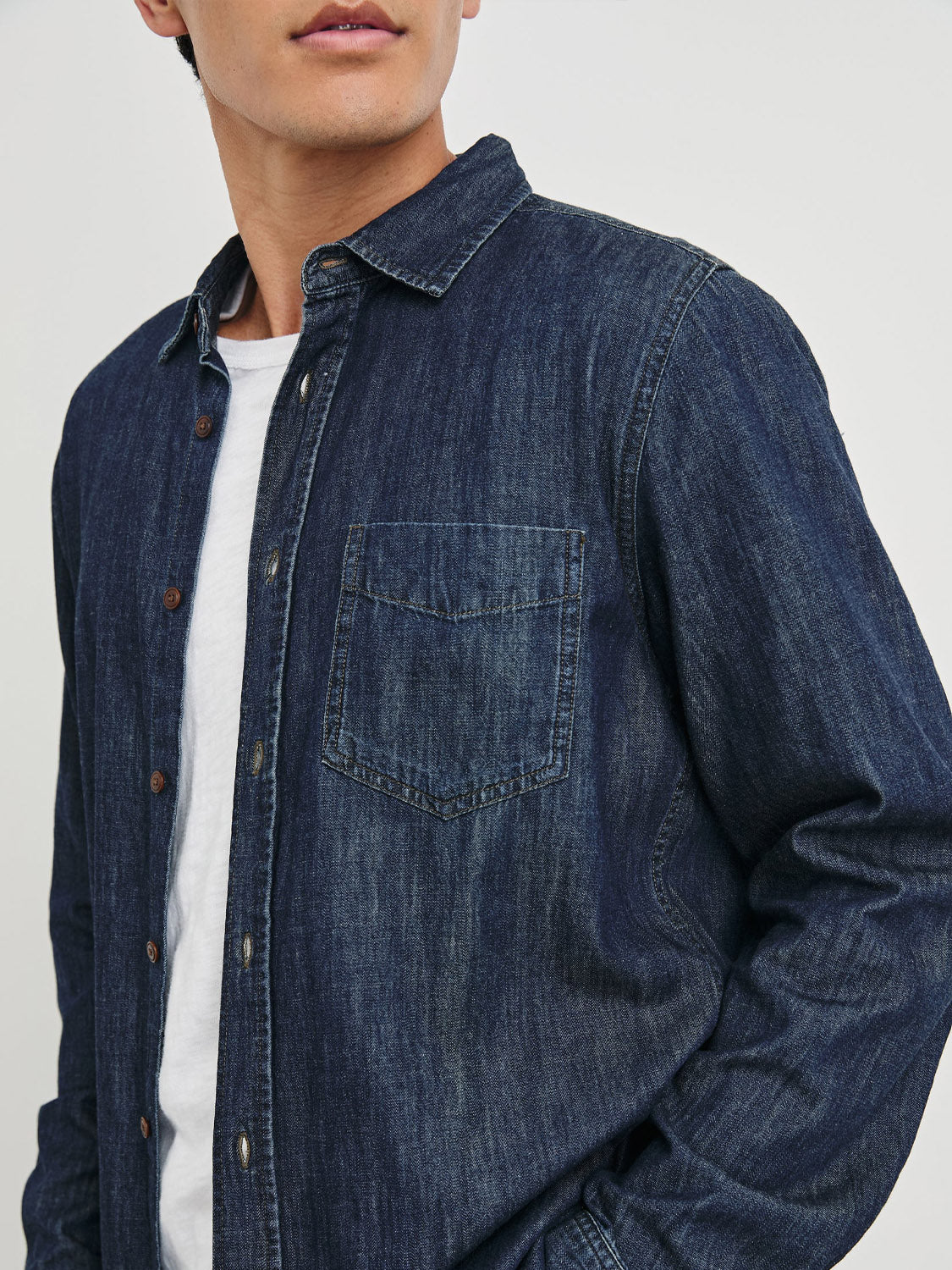 A person wearing the Rails Colton Dark Wash Indigo Shirt over a white T-shirt, showcasing its vintage-inspired denim texture and stitching. The background is plain and light-colored, focusing on the upper body.
